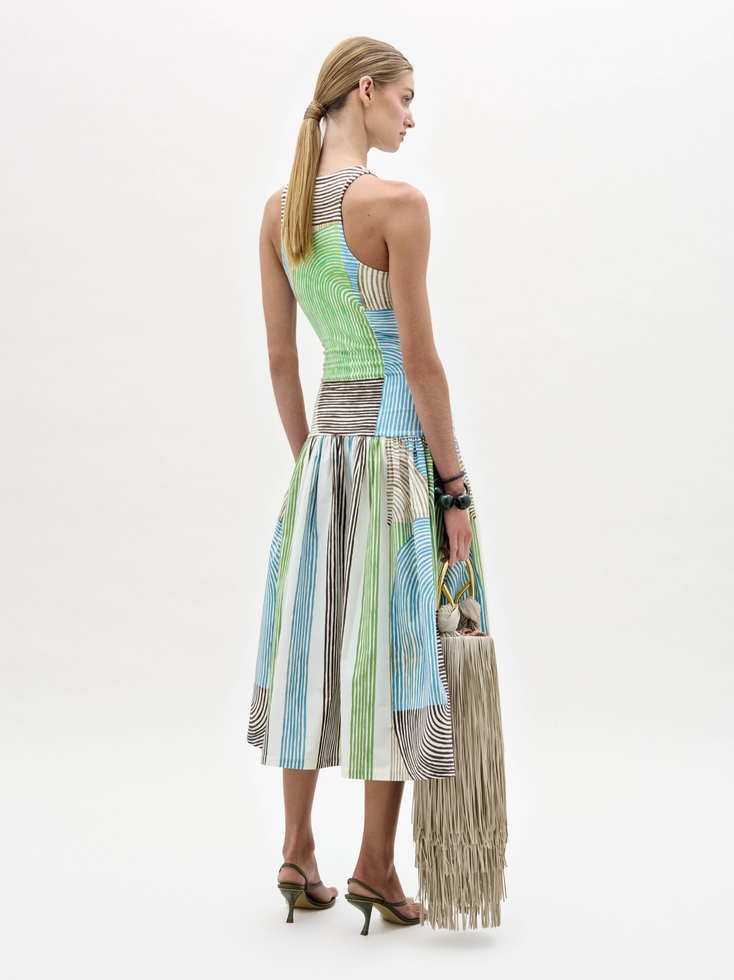 A woman models the Jamelia Dress, a sleeveless color-blocked midi with stripe patterns, styled with black heeled sandals and a fringed handbag, posing against a white background.
