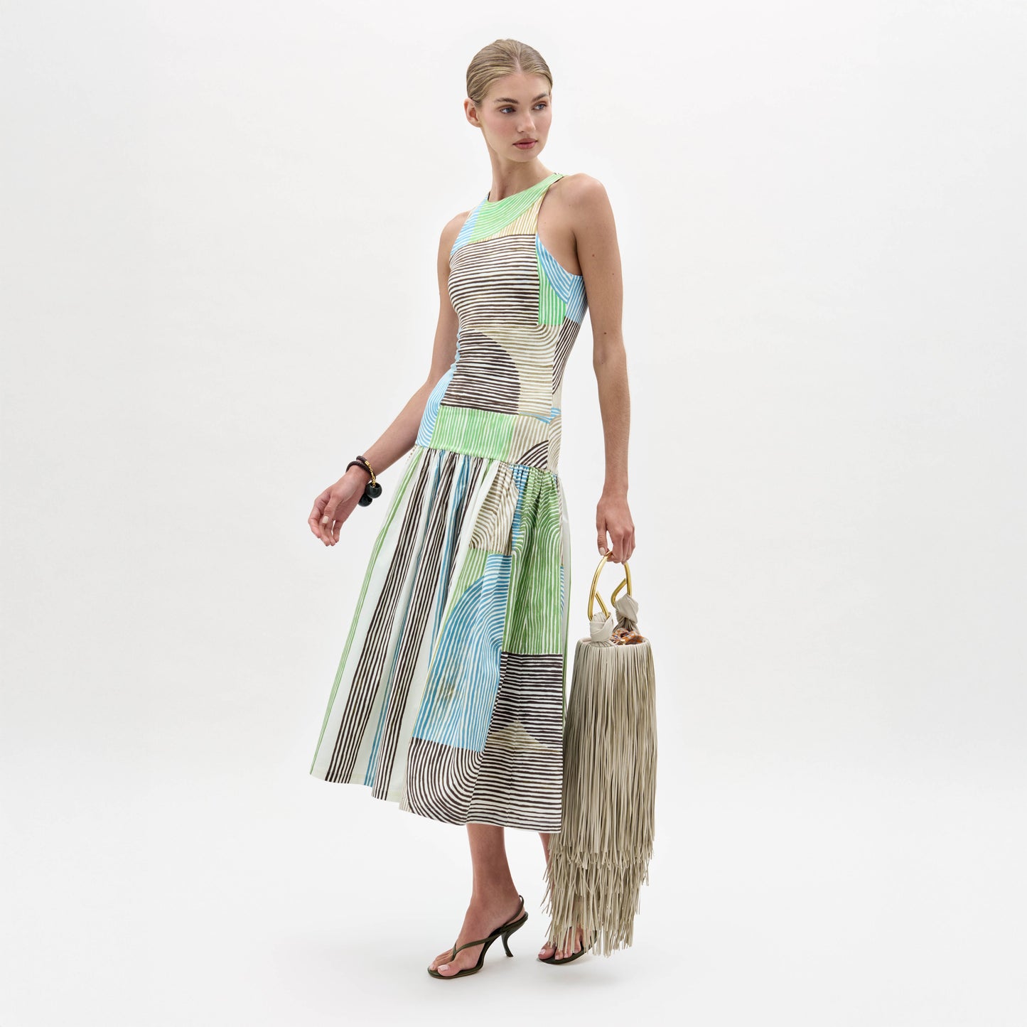 A woman models the Jamelia Dress, a sleeveless color-blocked midi with stripe patterns, styled with black heeled sandals and a fringed handbag, posing against a white background.