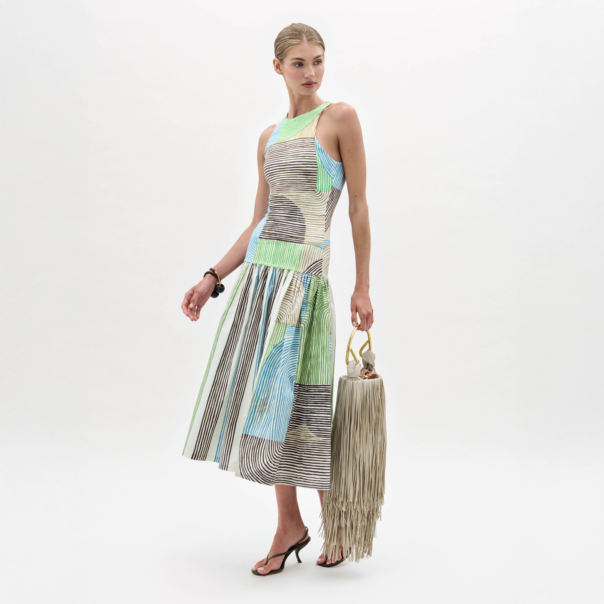 A woman models the Jamelia Dress, a sleeveless color-blocked midi with stripe patterns, styled with black heeled sandals and a fringed handbag, posing against a white background.