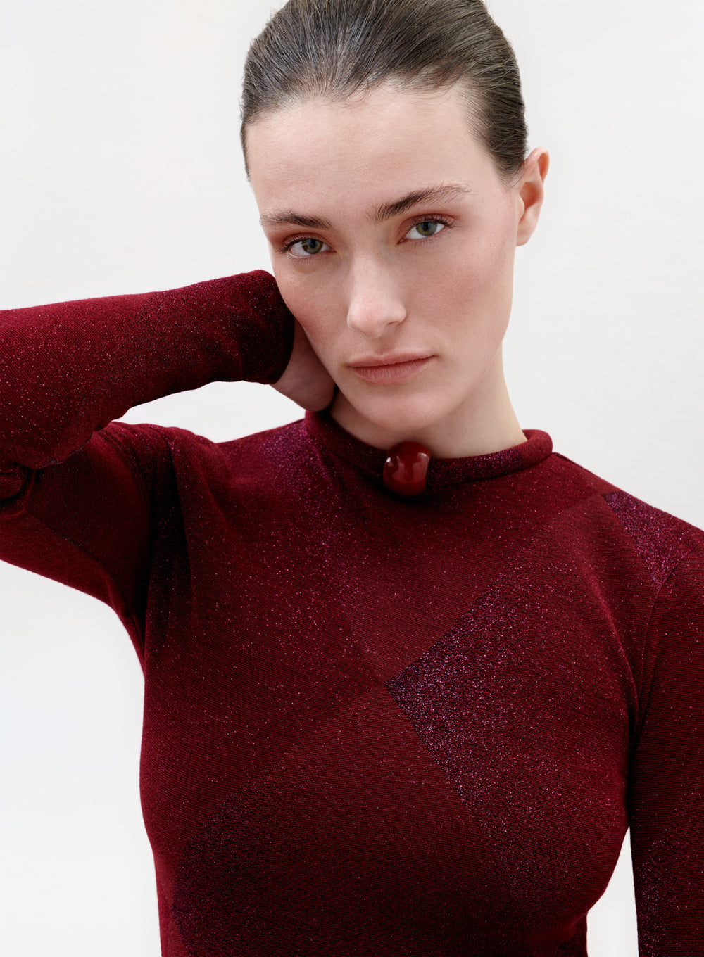 A woman with light skin and brown hair pulled back wears the Lalia T-Shirt in shimmering burgundy, rests one hand behind her neck, and looks directly at the camera with a neutral expression.