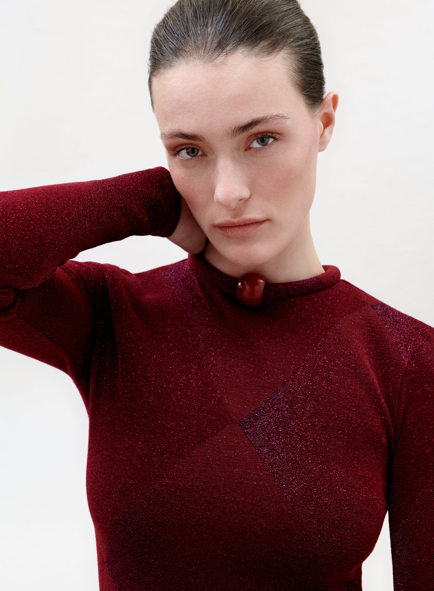 A woman with light skin and brown hair pulled back wears the Lalia T-Shirt in shimmering burgundy, rests one hand behind her neck, and looks directly at the camera with a neutral expression.