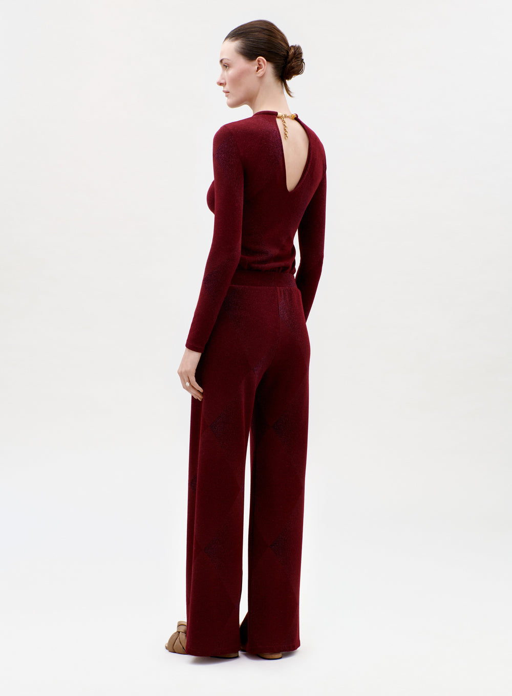 A woman stands facing away in the Colbie Pant and a maroon long-sleeve top with a back keyhole cutout. Her hair is in a neat bun, she wears brown shoes, and the background is plain and light.