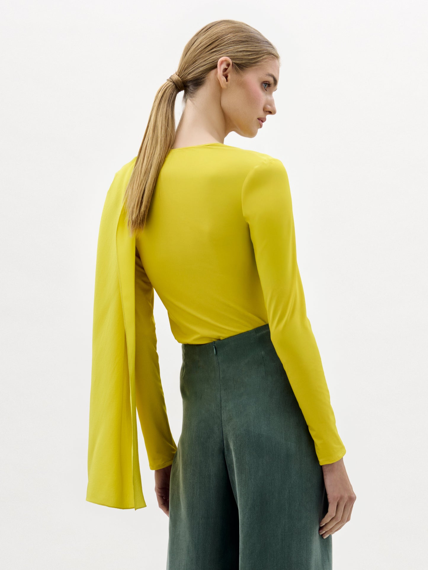 A woman stands against a plain background wearing the Josic Blouse, a yellow long-sleeve asymmetric top with a draped front, paired with a green skirt tied at the waist.