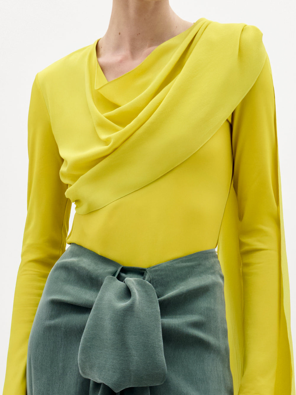 A woman stands against a plain background wearing the Josic Blouse, a yellow long-sleeve asymmetric top with a draped front, paired with a green skirt tied at the waist.
