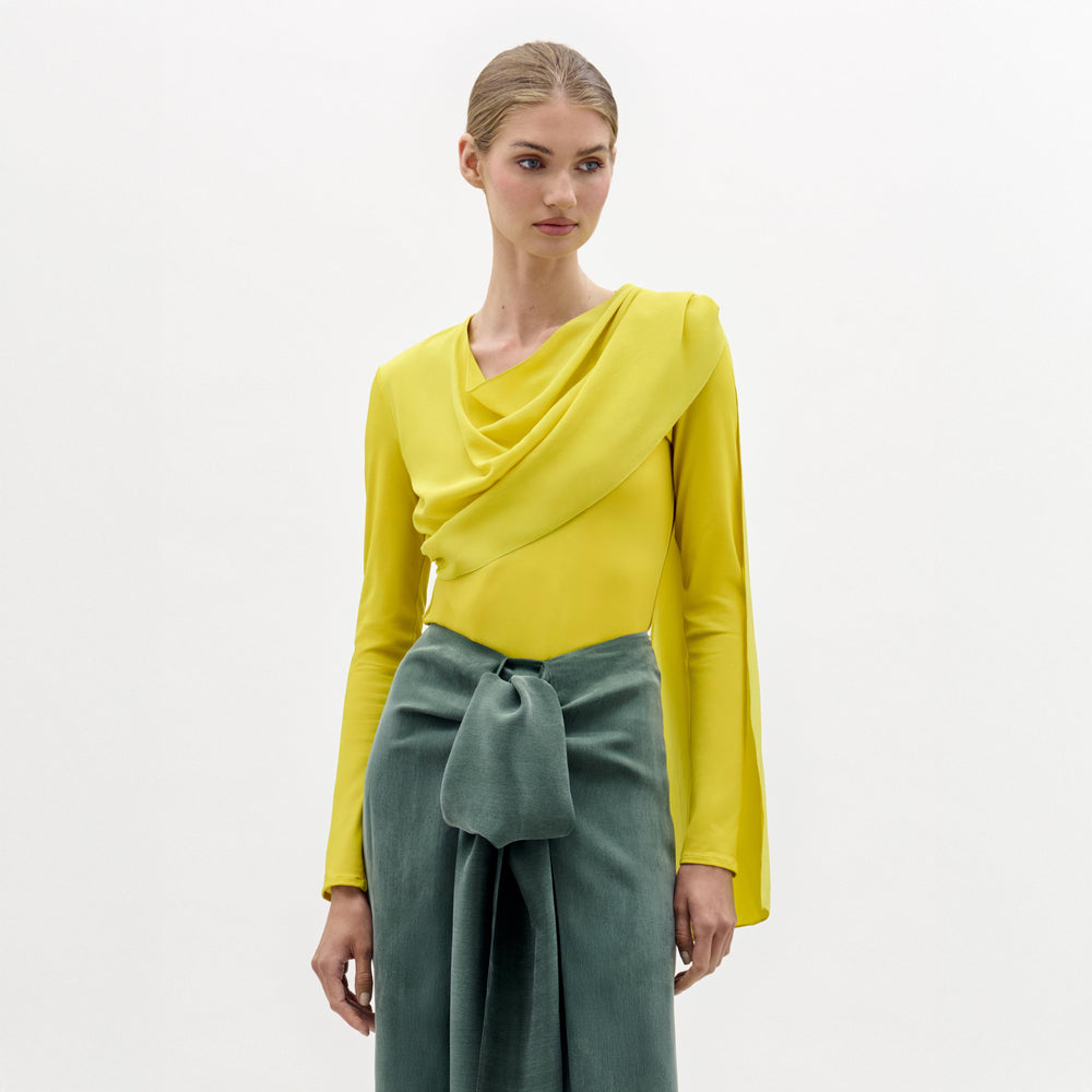 A woman stands against a plain background wearing the Josic Blouse, a yellow long-sleeve asymmetric top with a draped front, paired with a green skirt tied at the waist.