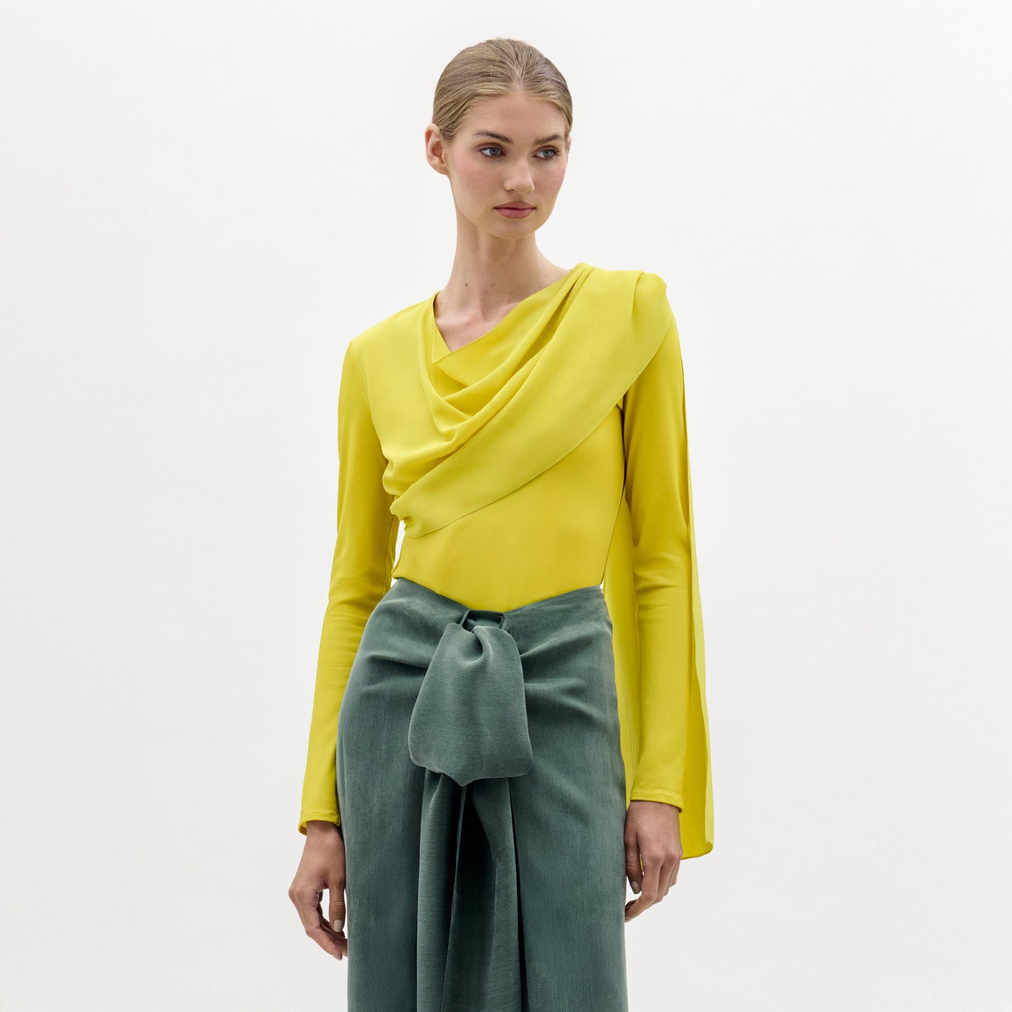 A woman stands against a plain background wearing the Josic Blouse, a yellow long-sleeve asymmetric top with a draped front, paired with a green skirt tied at the waist.