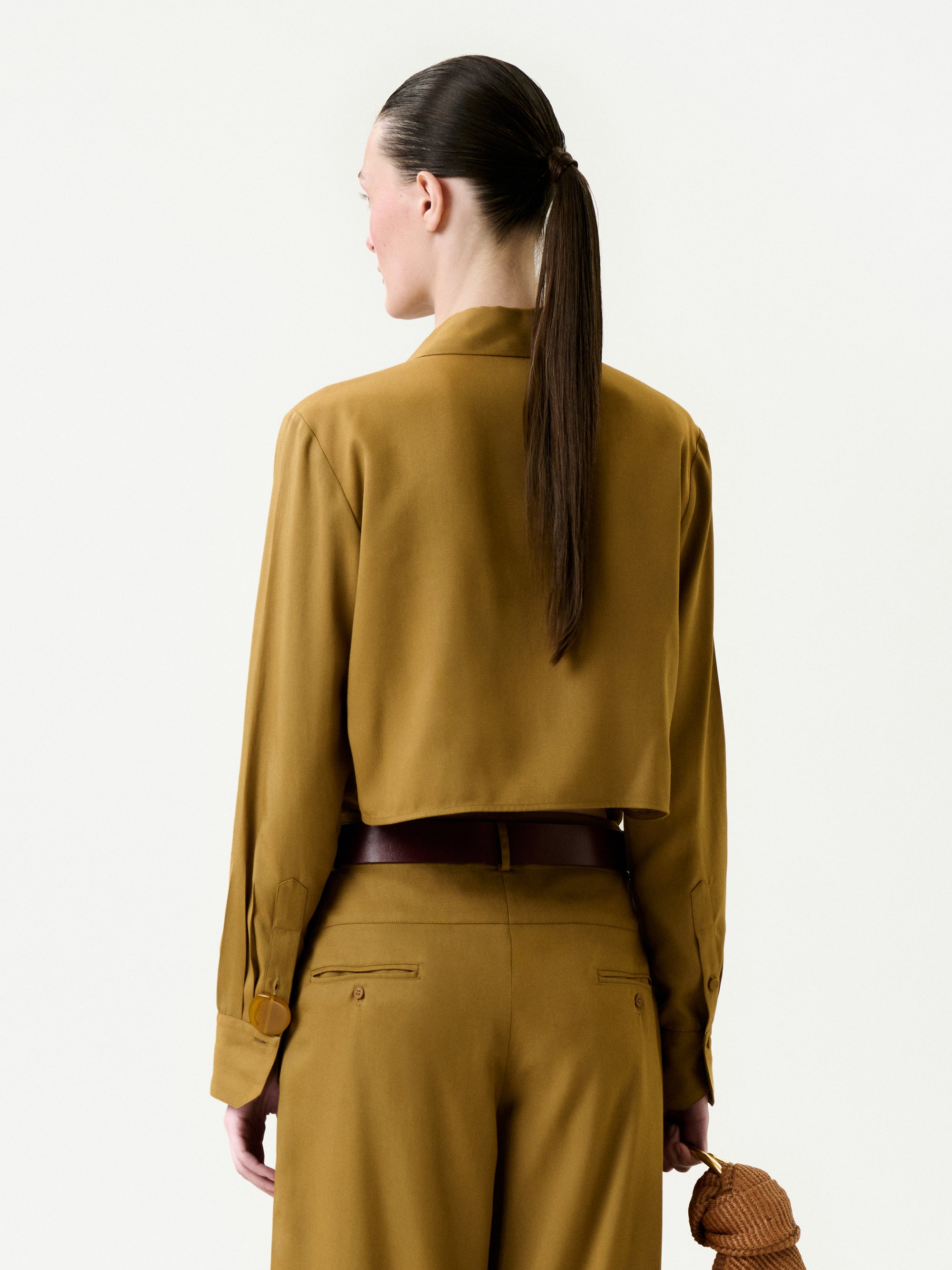 A person with long brown hair in a ponytail is shown from behind, wearing the mustard-colored Mazz Blouse with matching trousers and holding a small brown textured clutch bag in their right hand.