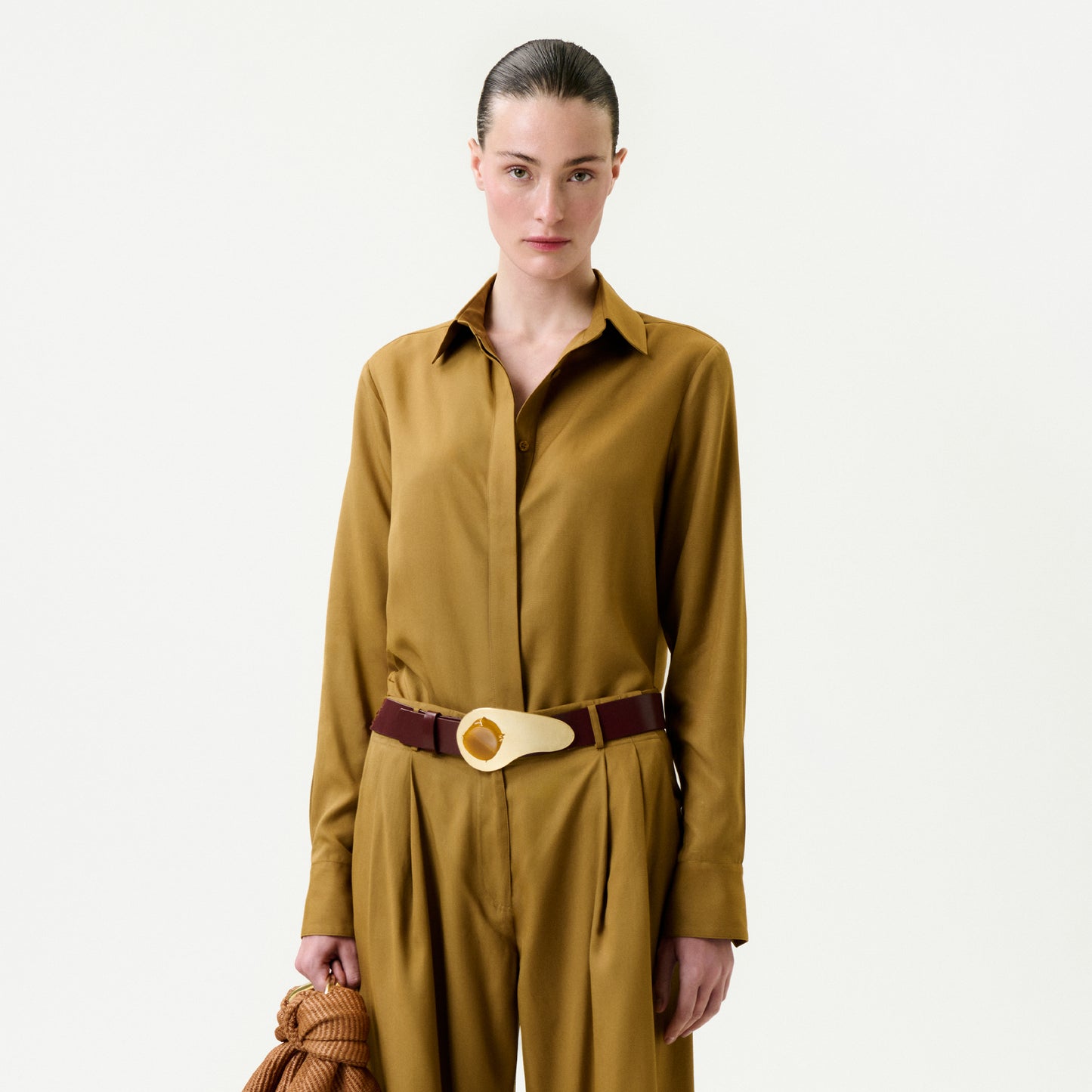 A woman with dark hair pulled back wears the Mazz Blouse in mustard yellow and matching wide-leg 100% Lyocell trousers, accessorized with a brown belt with a large gold buckle. She holds a textured brown item in her left hand against a light background.