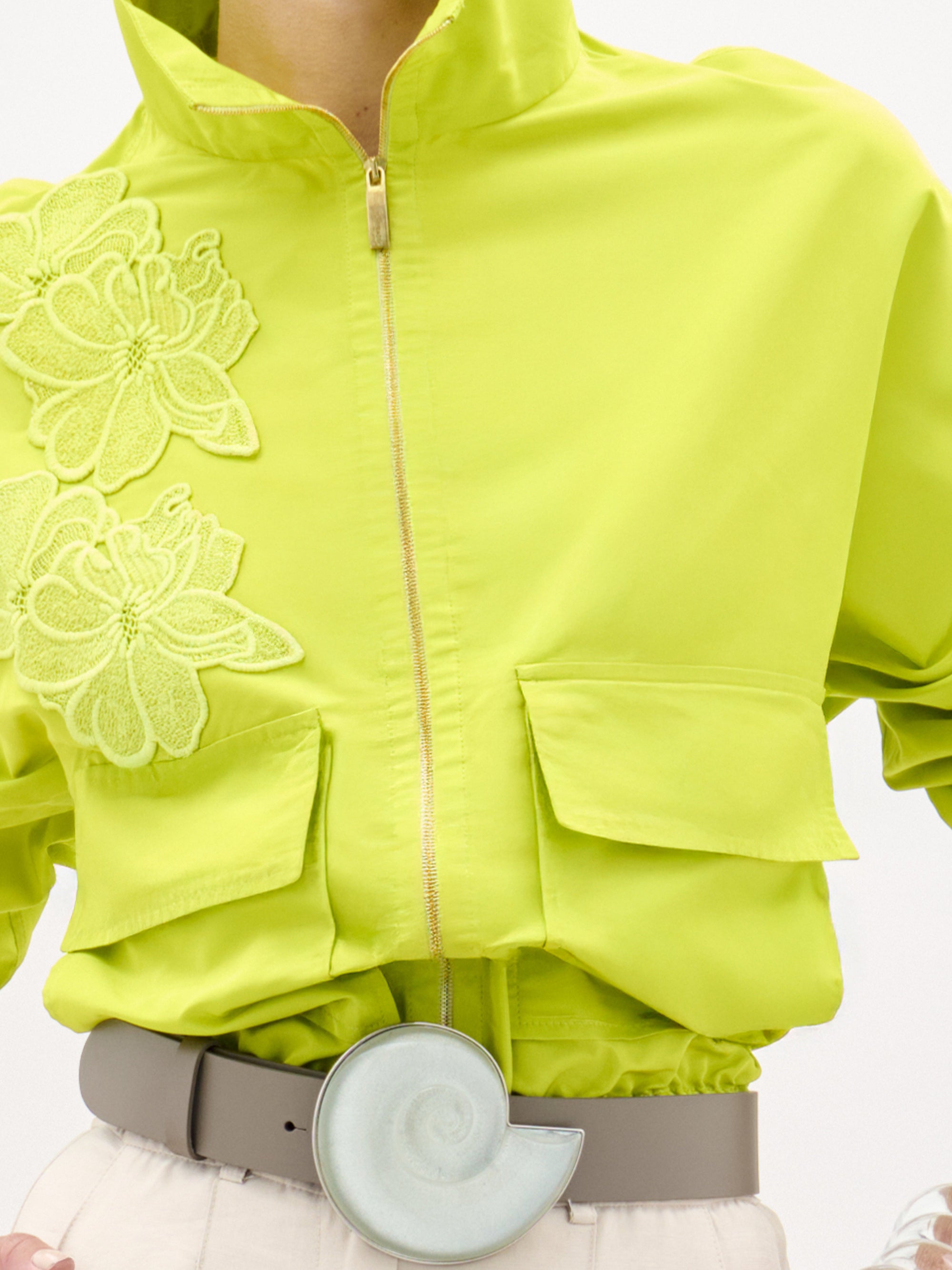 A woman wears the Donata Jacket, a bright yellow cropped jacket with floral embroidery, paired with beige high-waisted pants, a wide belt featuring a circular buckle, and a clear bracelet as she stands against a plain background.