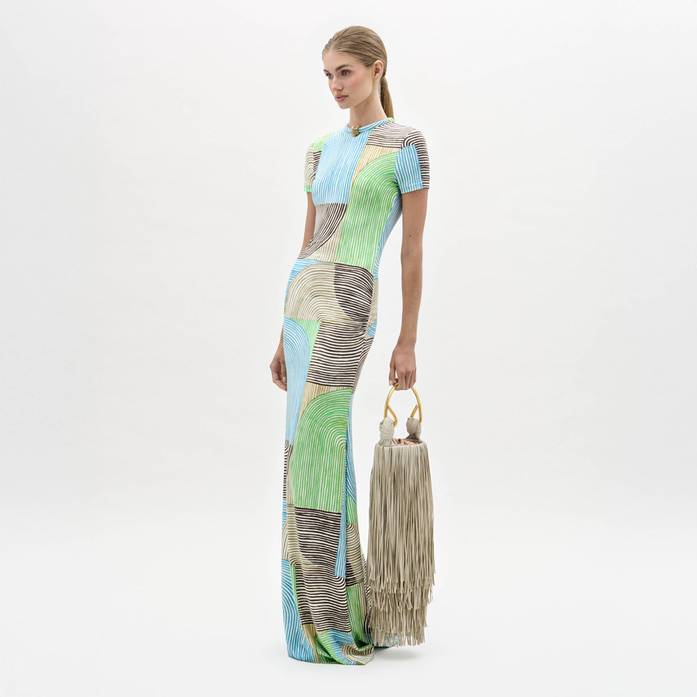 A woman in a Yara Dress, featuring multicolored patterns, stands holding a large beige fringe bag with circular handles against a plain white background.