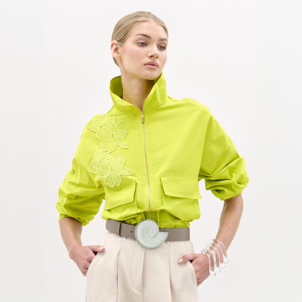A woman wears the Donata Jacket, a bright yellow cropped jacket with floral embroidery, paired with beige high-waisted pants, a wide belt featuring a circular buckle, and a clear bracelet as she stands against a plain background.