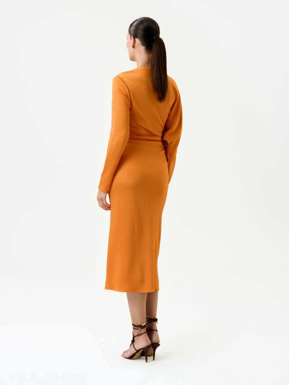 A woman with dark, straight hair in a low ponytail wears the Shiloh Dress—a long-sleeved, orange midi dress made from luxurious Italian fabric—with brown strappy heels, standing against a plain white background.