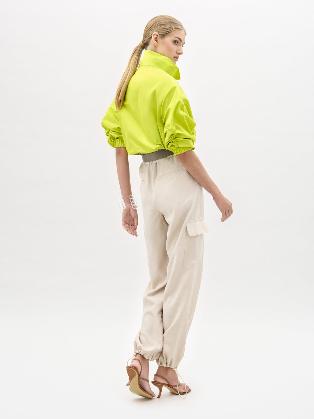 A woman stands against a plain white background wearing a bright green cropped jacket, the Darnell Pant in high-waist beige Italian fabric, a wide white belt, and heeled sandals.