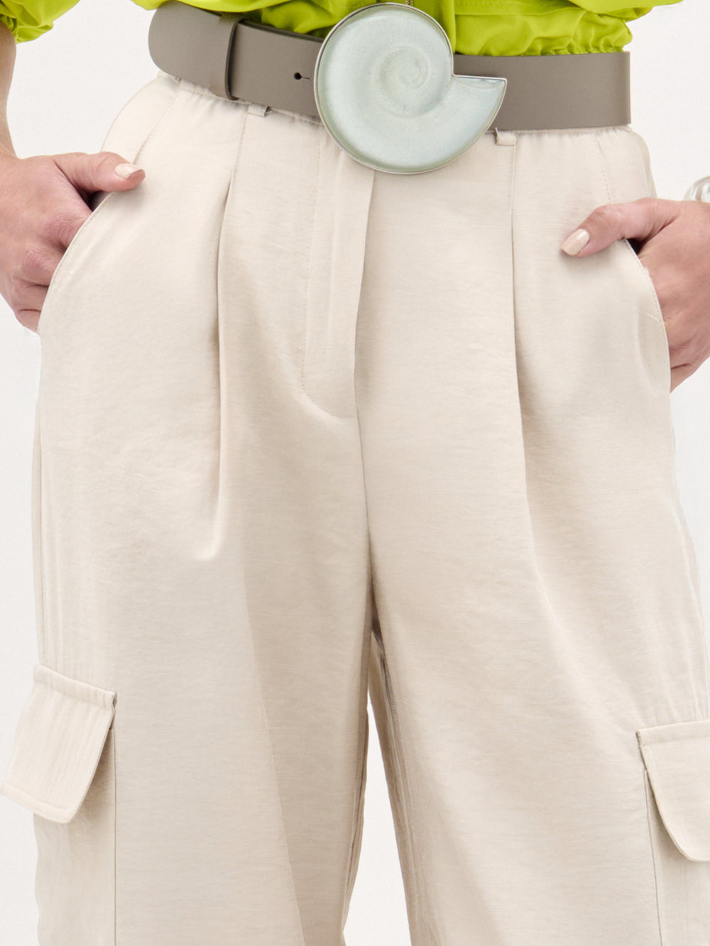 A woman stands against a plain white background wearing a bright green cropped jacket, the Darnell Pant in high-waist beige Italian fabric, a wide white belt, and heeled sandals.