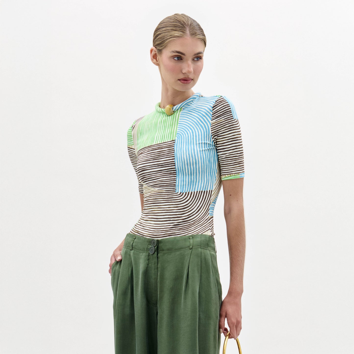 A woman stands against a plain background wearing the Varinia Tshirt with stripes and loose-fitting green trousers, holding a small yellow bag.
