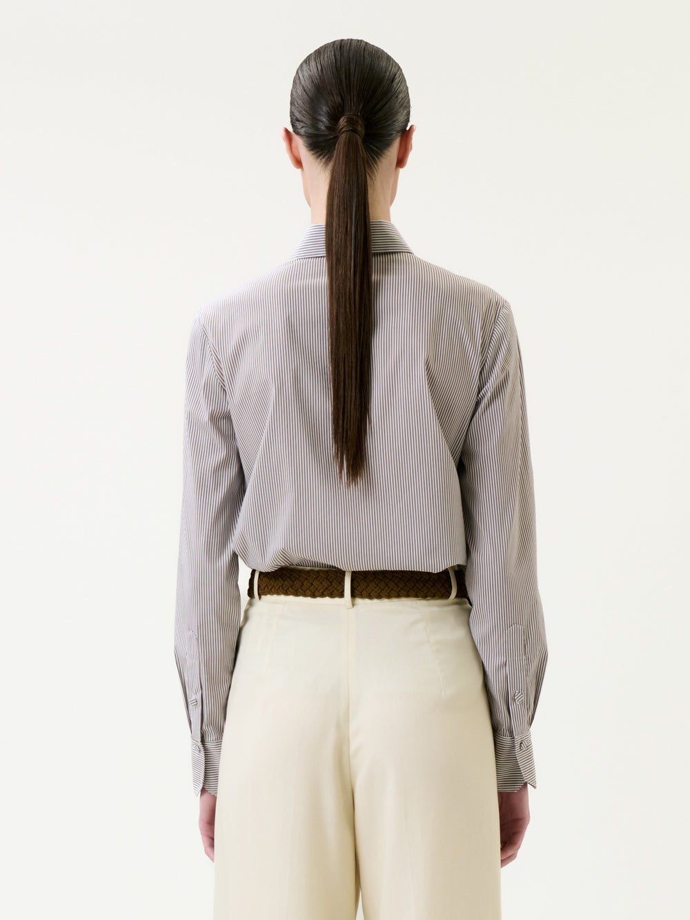 A person stands with their back to the camera, wearing the Warda Shirt—a striped button-down—tucked into high-waisted, light-colored pants. Their long, straight hair is tied in a low ponytail. The background is plain white.