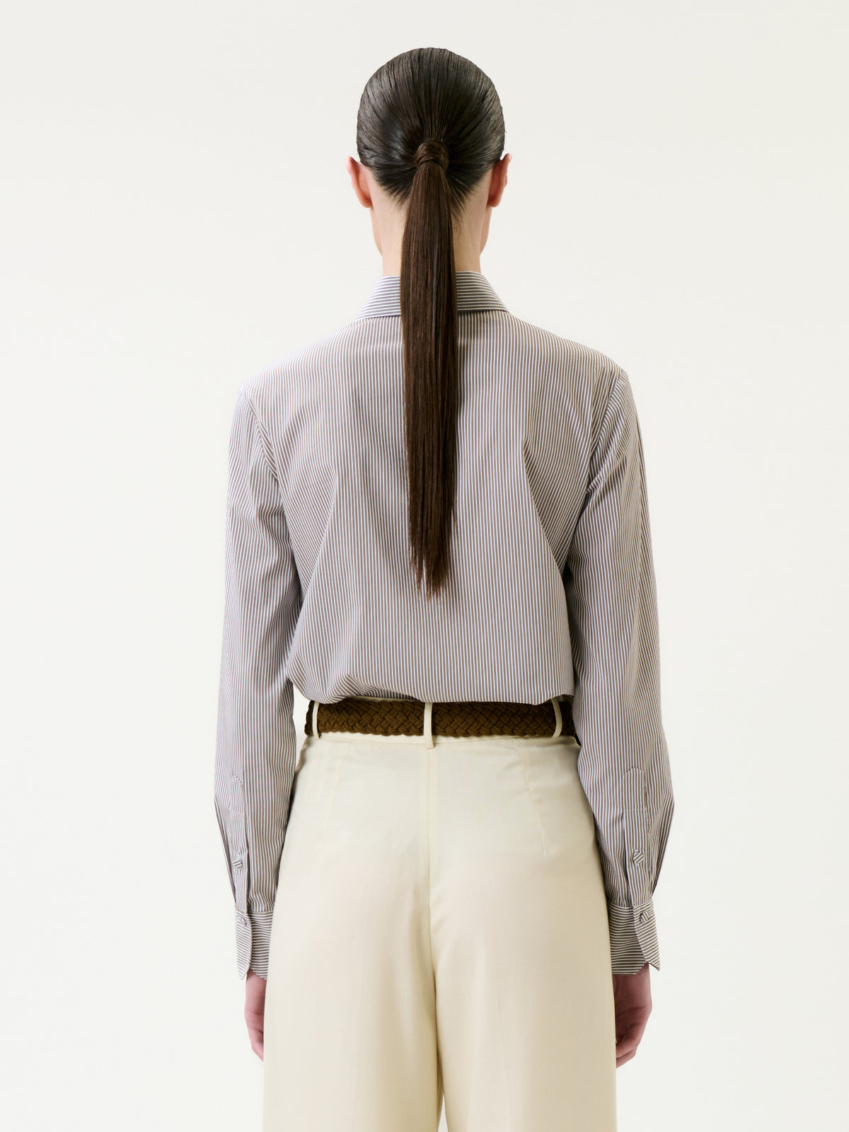 A person stands with their back to the camera, wearing the Warda Shirt—a striped button-down—tucked into high-waisted, light-colored pants. Their long, straight hair is tied in a low ponytail. The background is plain white.