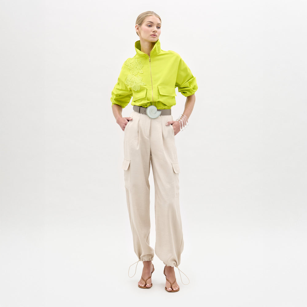 A woman stands against a plain white background wearing a bright green cropped jacket, the Darnell Pant in high-waist beige Italian fabric, a wide white belt, and heeled sandals.