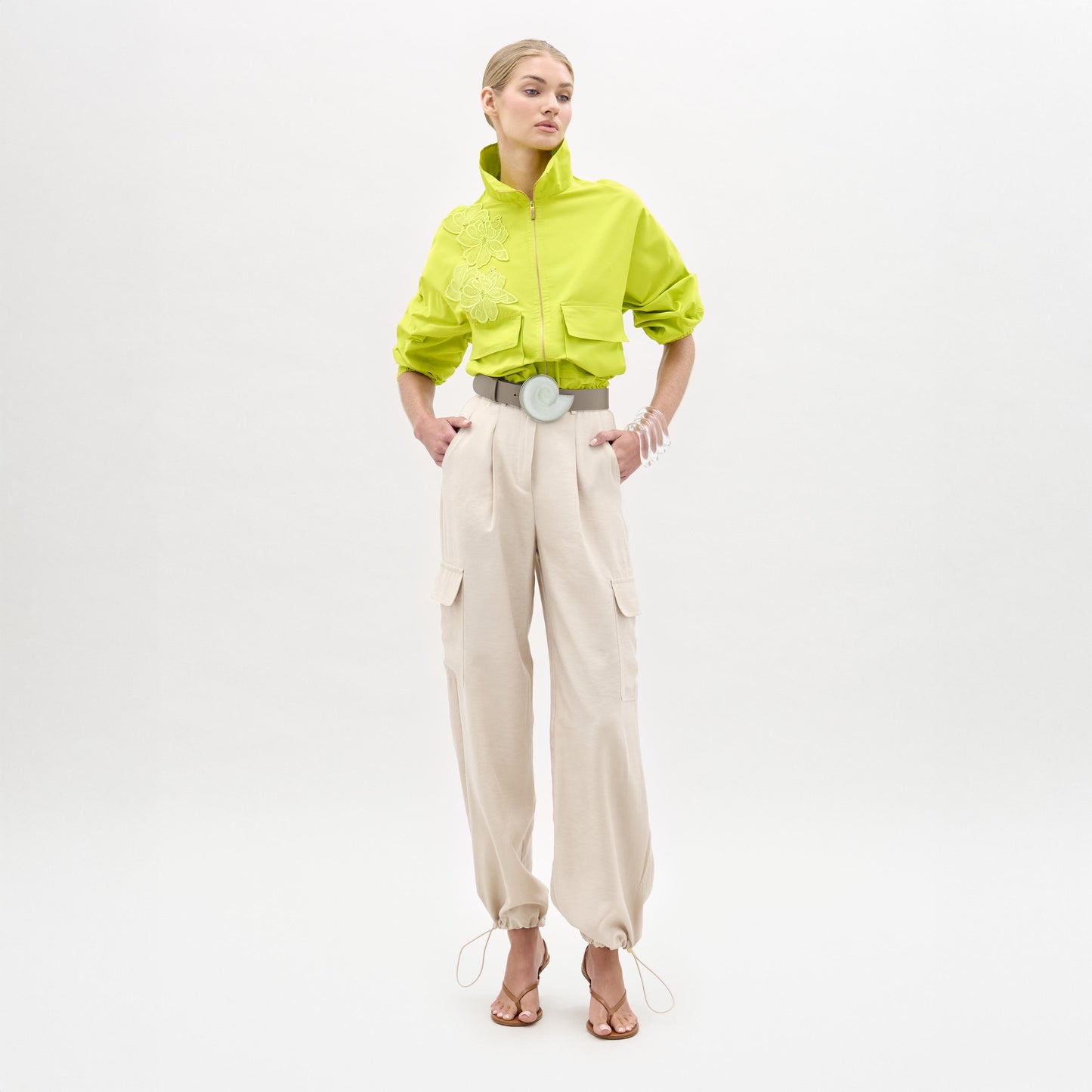 A woman stands against a plain white background wearing a bright green cropped jacket, the Darnell Pant in high-waist beige Italian fabric, a wide white belt, and heeled sandals.
