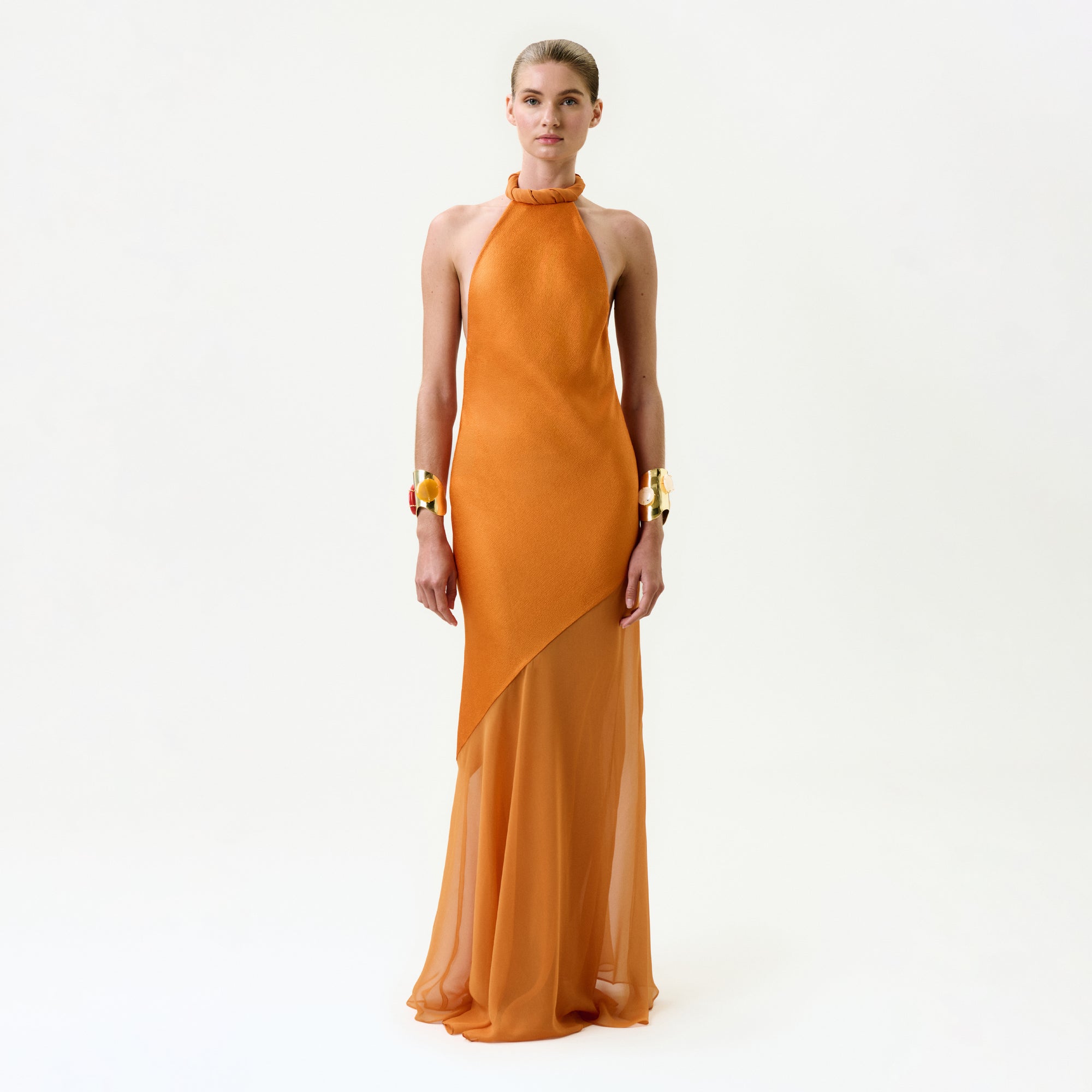 A person stands against a plain white background wearing the Keisha Dress—a sleeveless, bright orange silk halter-neck maxi with an asymmetrical sheer skirt—accessorized with bold gold bracelets on both wrists.