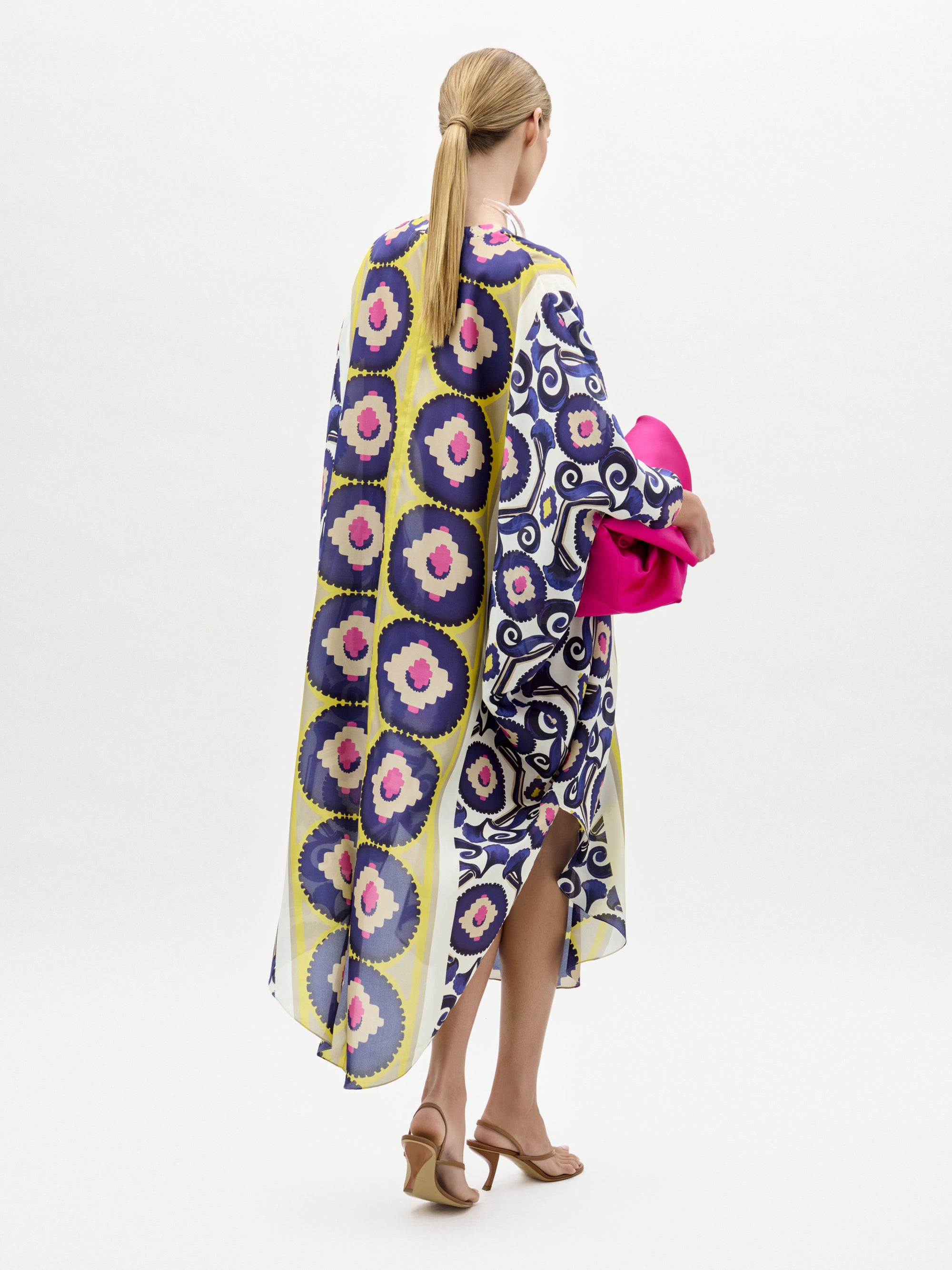 Woman models the Ilse Tunic, a long maxi dress crafted from Italian fabric featuring blue, yellow, and pink circles. She holds a bright pink clutch and wears heeled sandals against a plain white background.