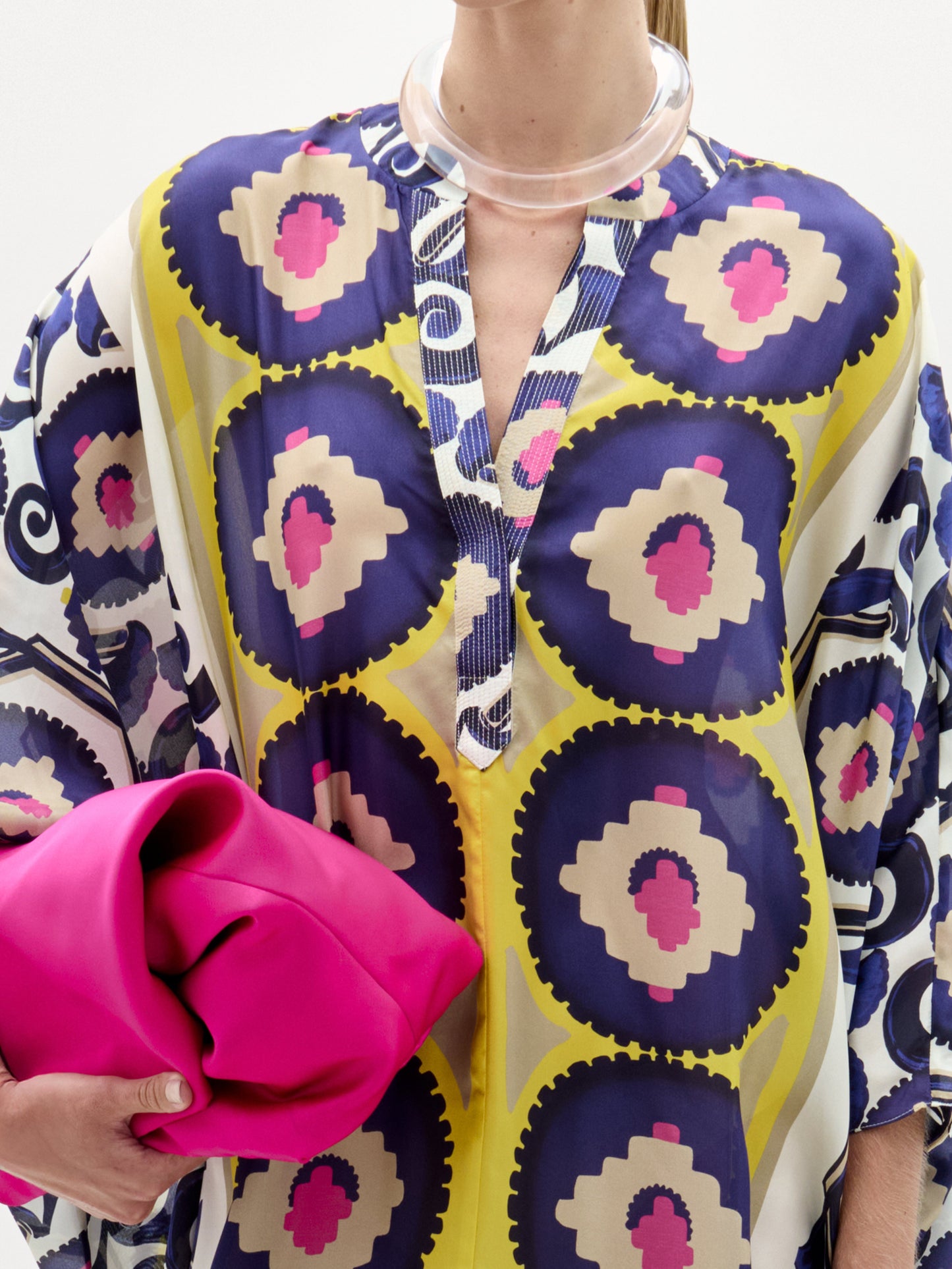 Woman models the Ilse Tunic, a long maxi dress crafted from Italian fabric featuring blue, yellow, and pink circles. She holds a bright pink clutch and wears heeled sandals against a plain white background.