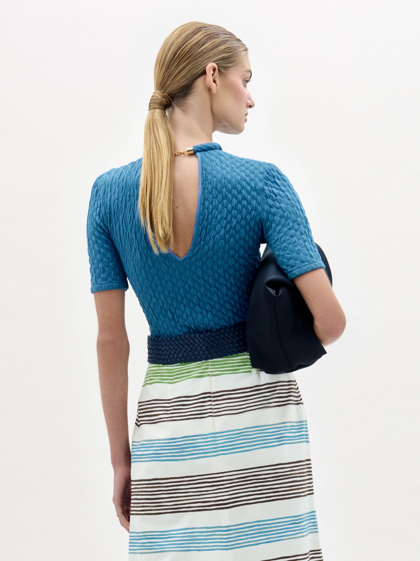 A woman in a blue Venecia Tshirt and striped skirt holds a black clutch. Her straight hair is pulled back, and she wears a large oval belt buckle.