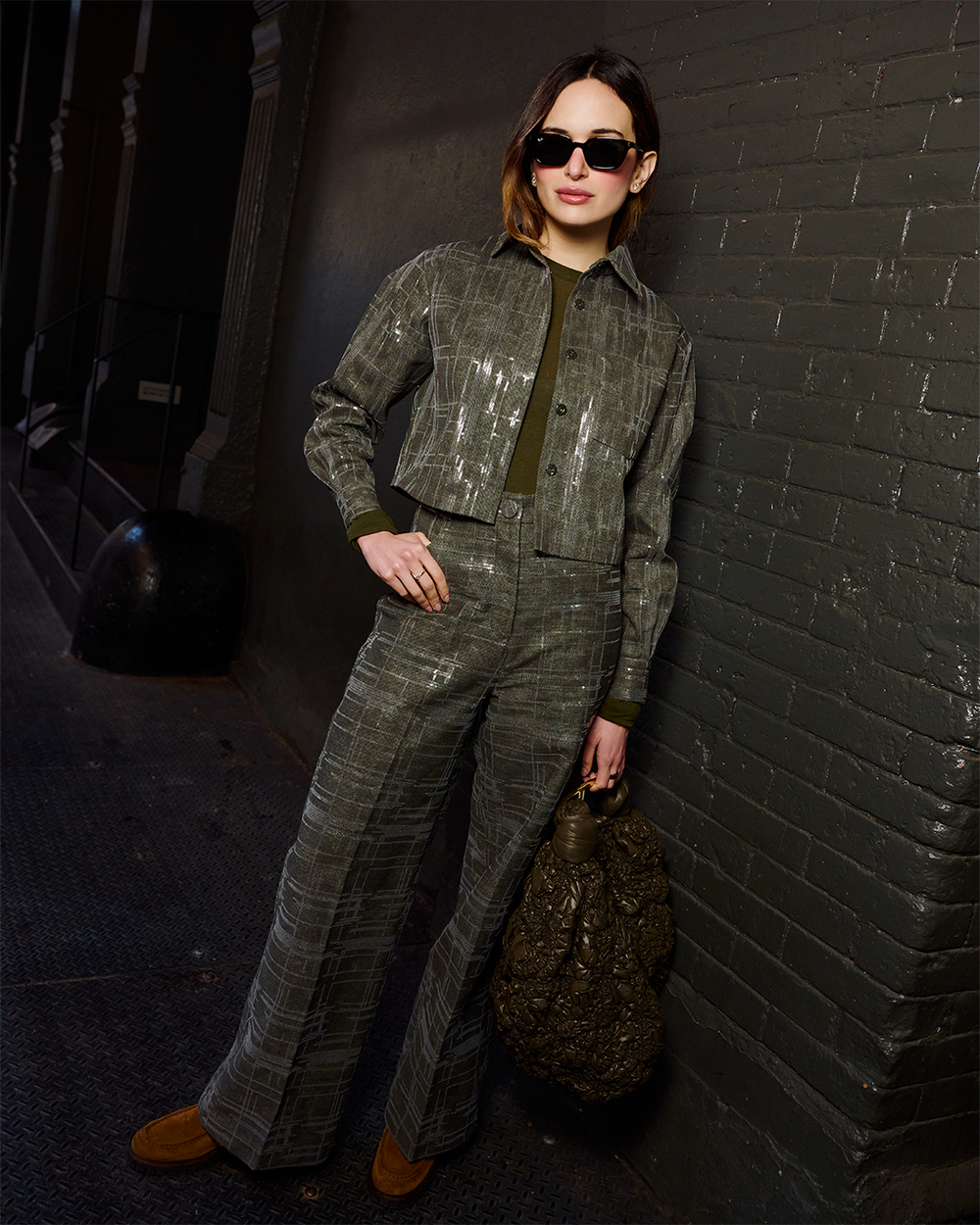 A woman stands against a dark brick wall wearing a matching gray plaid cropped jacket and wide-leg trousers set with olive green trim, black sunglasses, and brown suede boots, while holding a textured dark brown bag.