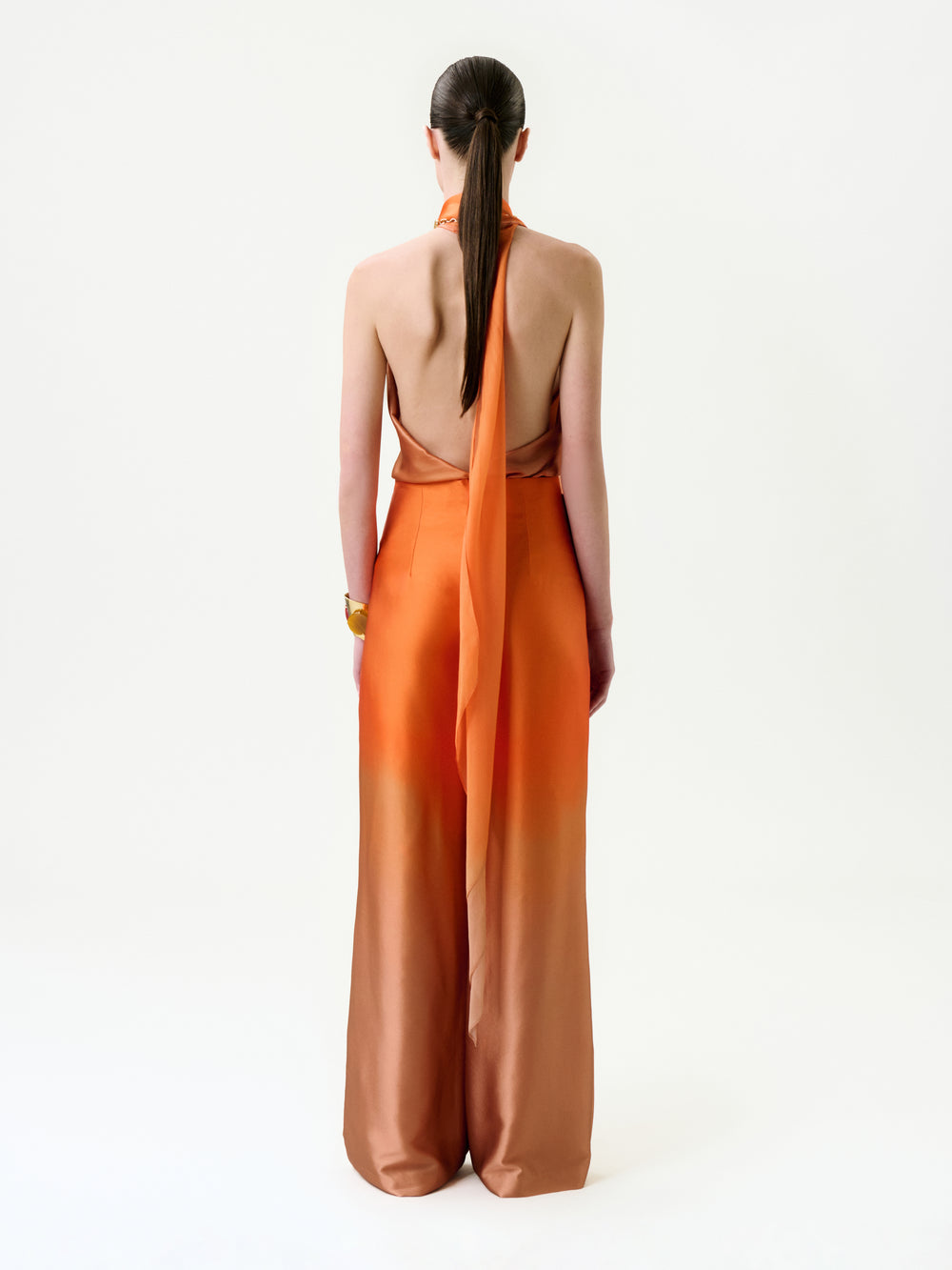 A woman with straight brown hair in a low ponytail is shown from behind, wearing the Andie Pant in orange Italian fabric, featuring a halter neck, long sash, wide legs, and a gold bracelet on her right wrist.