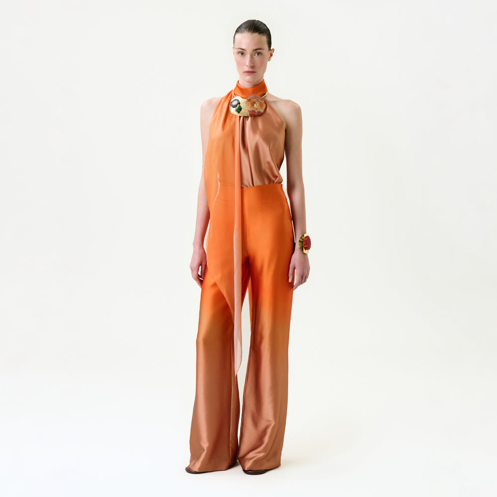 A person poses against a white background, wearing the Andie Pant—a chic sleeveless orange jumpsuit with flared legs, sheer neck scarf detail, and bold statement jewelry. Crafted from luxurious Italian fabric.
