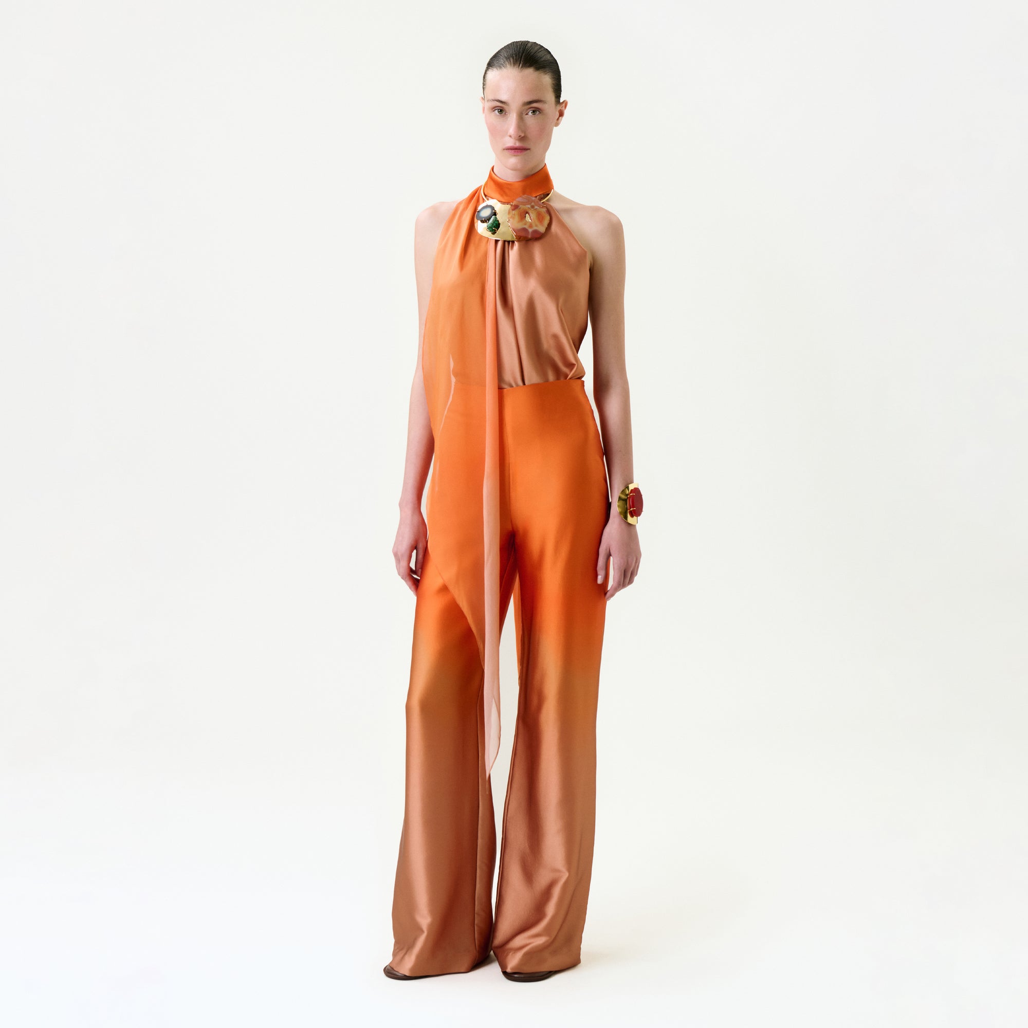A person poses against a white background, wearing the Andie Pant—a chic sleeveless orange jumpsuit with flared legs, sheer neck scarf detail, and bold statement jewelry. Crafted from luxurious Italian fabric.