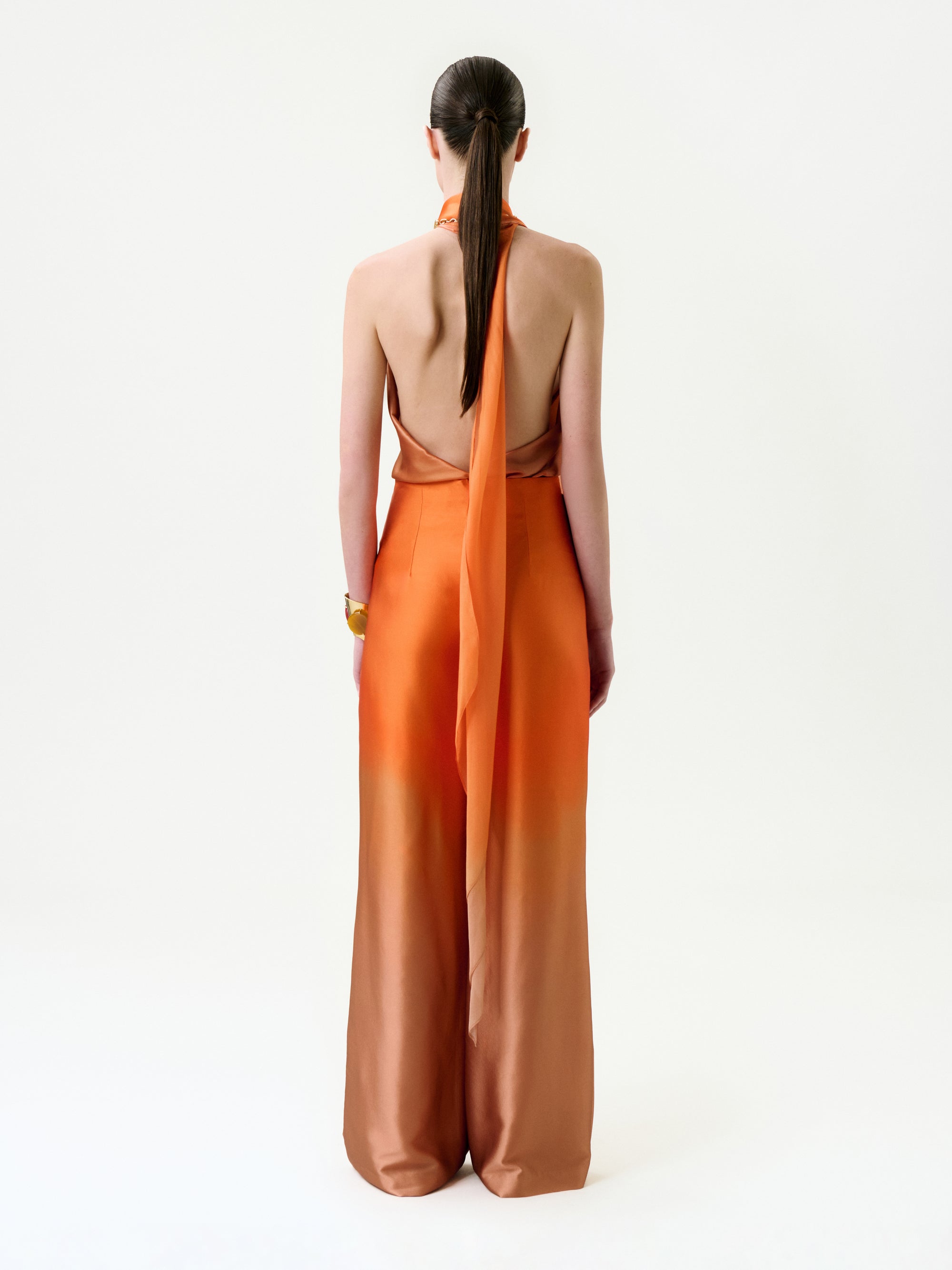 A woman with straight brown hair in a low ponytail wears the Velia Blouse, crafted from luxurious Italian fabric in an orange ombre hue, standing against a plain white background and facing away from the camera.