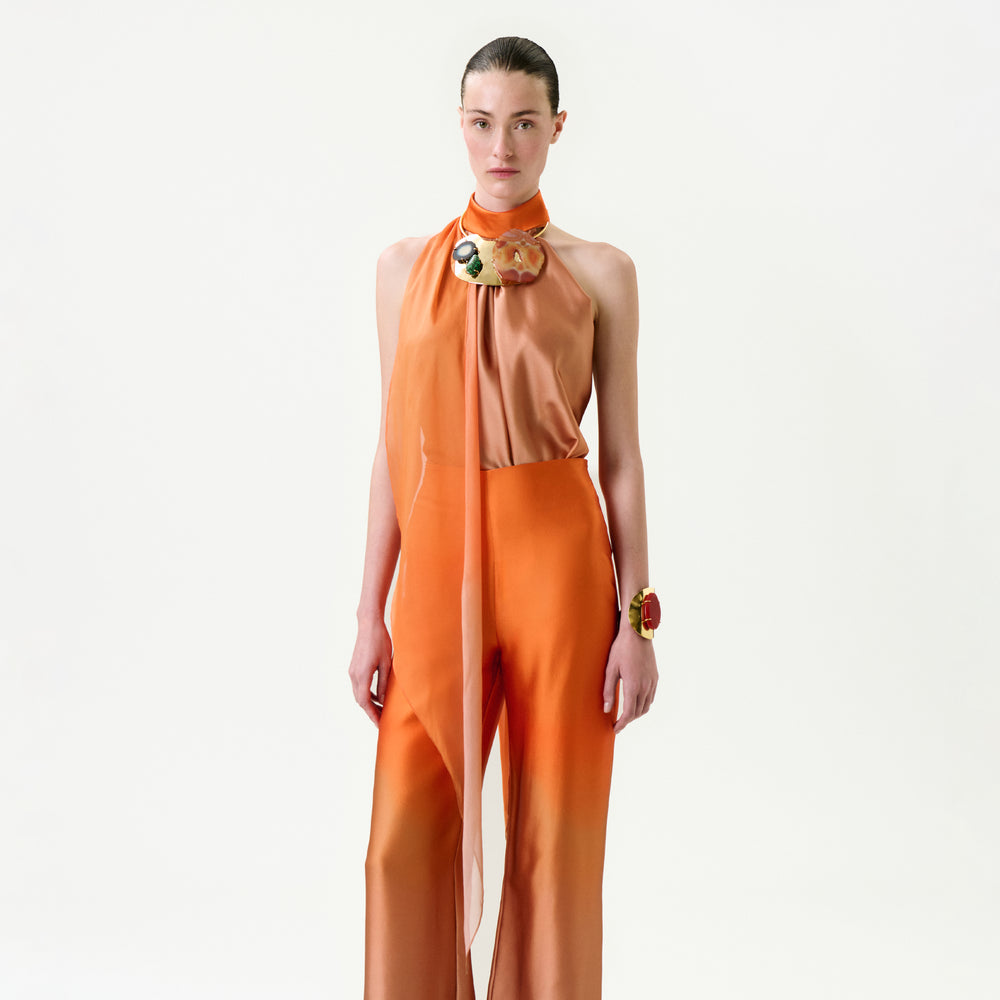 A woman stands against a white background wearing the Velia Blouse—an elegant sleeveless orange top with a high neck and sheer scarf detail, crafted from Italian fabric. She has slicked-back hair with a bold bracelet and brooch.