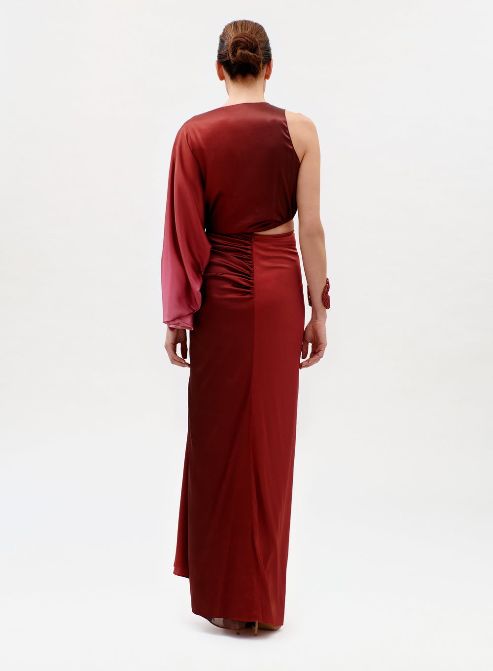 A woman with her hair in a neat bun stands facing away, wearing the Elimar Dress—a floor-length dark red gown with a single long sleeve on the left arm—against a plain light-colored background.
