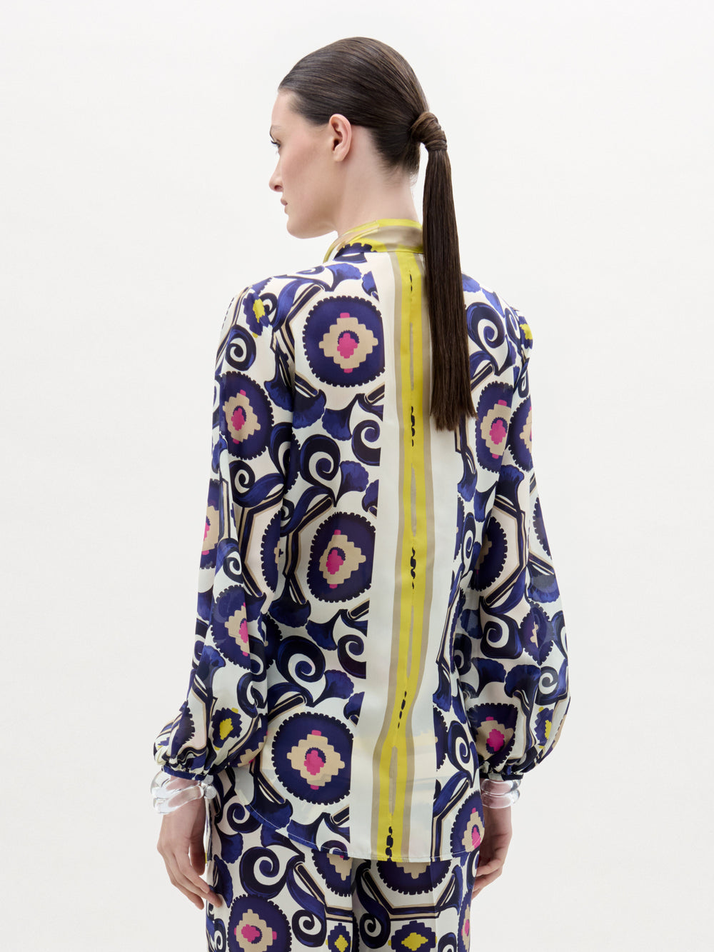 A woman stands against a white background wearing the Honey Blouse, a patterned silk long-sleeve top with a front twist detail and vibrant yellow, purple, blue, and pink circular and geometric designs.