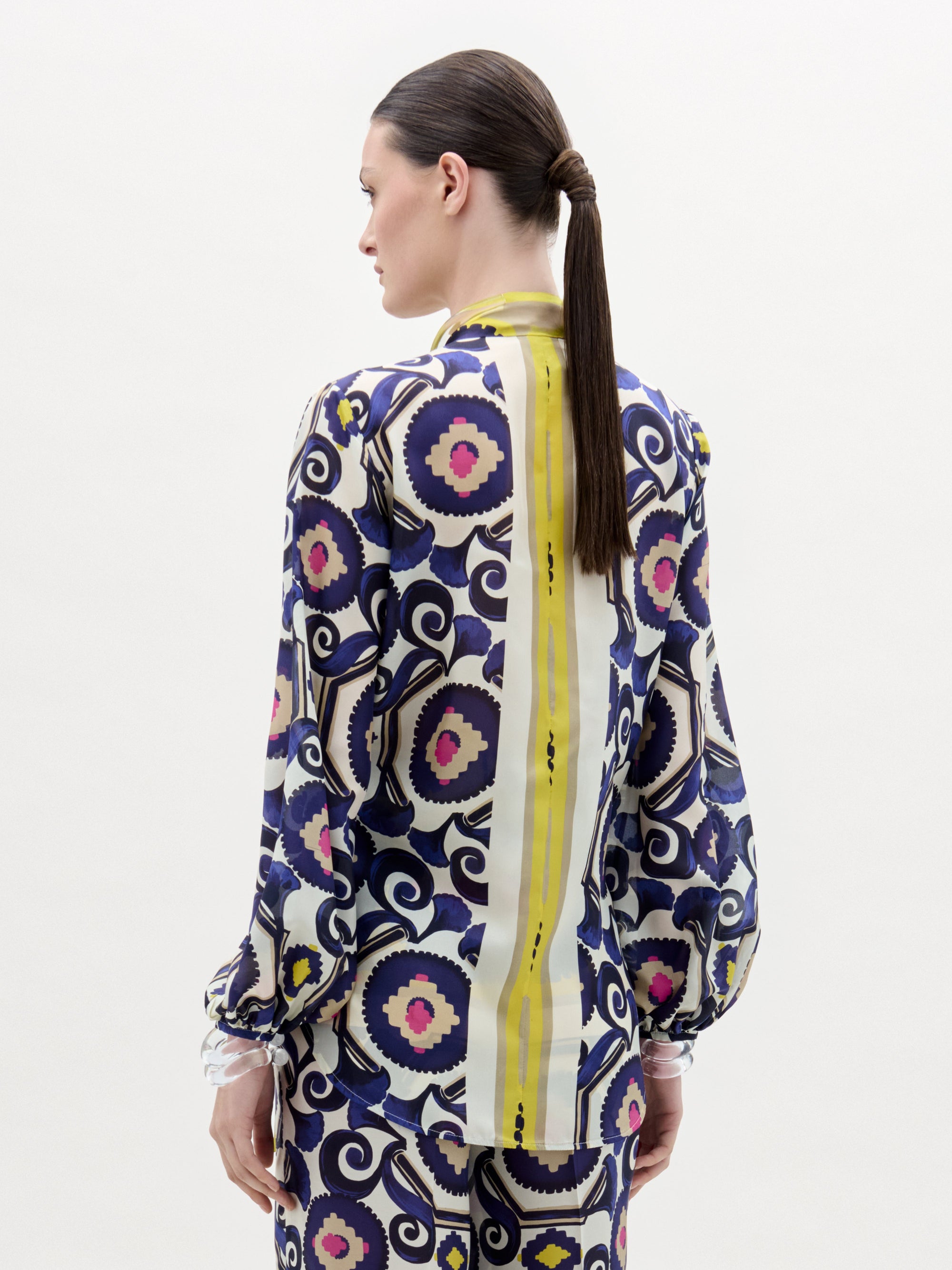 A woman stands against a white background wearing the Honey Blouse, a patterned silk long-sleeve top with a front twist detail and vibrant yellow, purple, blue, and pink circular and geometric designs.