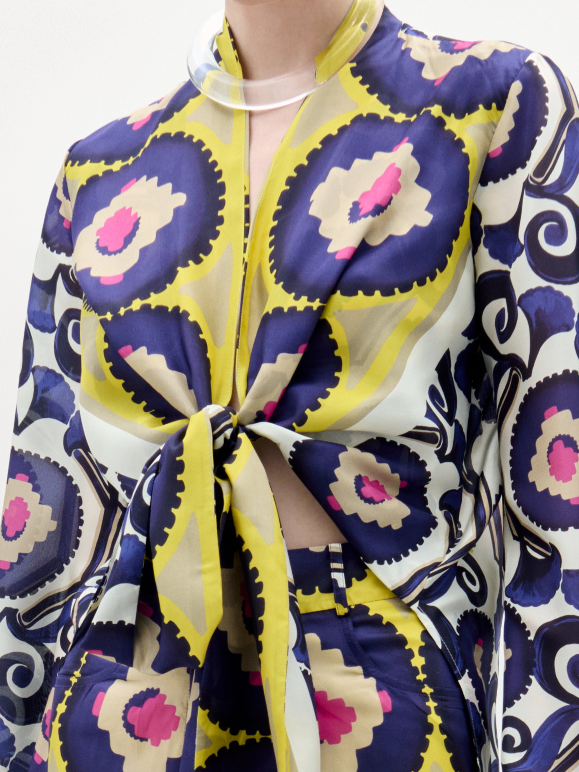A woman stands against a white background wearing the Honey Blouse, a patterned silk long-sleeve top with a front twist detail and vibrant yellow, purple, blue, and pink circular and geometric designs.