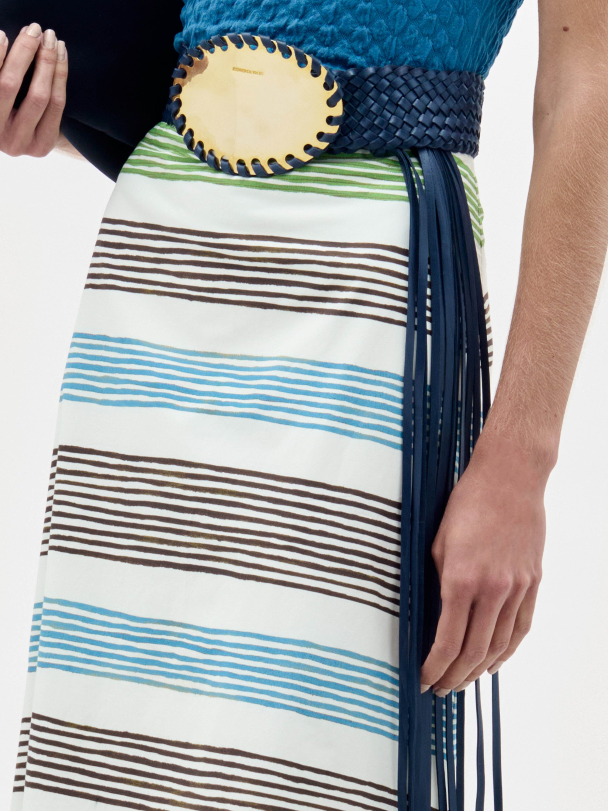 A woman stands against a plain white background wearing the Lyla Skirt with geometric stripes, a blue textured top, wide belt with a large gold buckle, heeled sandals, and holding a dark clutch.