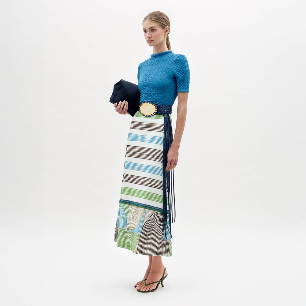 A woman stands against a plain white background wearing the Lyla Skirt with geometric stripes, a blue textured top, wide belt with a large gold buckle, heeled sandals, and holding a dark clutch.
