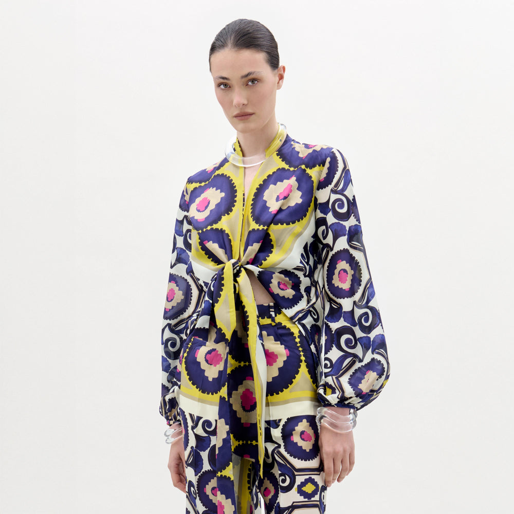 A woman stands against a white background wearing the Honey Blouse, a patterned silk long-sleeve top with a front twist detail and vibrant yellow, purple, blue, and pink circular and geometric designs.