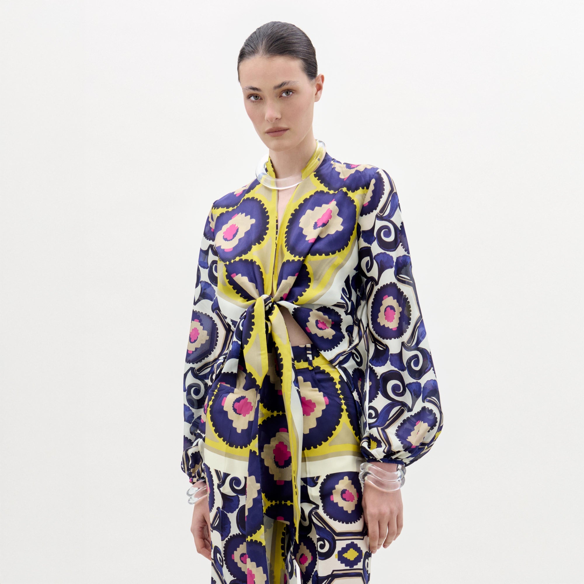 A woman stands against a white background wearing the Honey Blouse, a patterned silk long-sleeve top with a front twist detail and vibrant yellow, purple, blue, and pink circular and geometric designs.