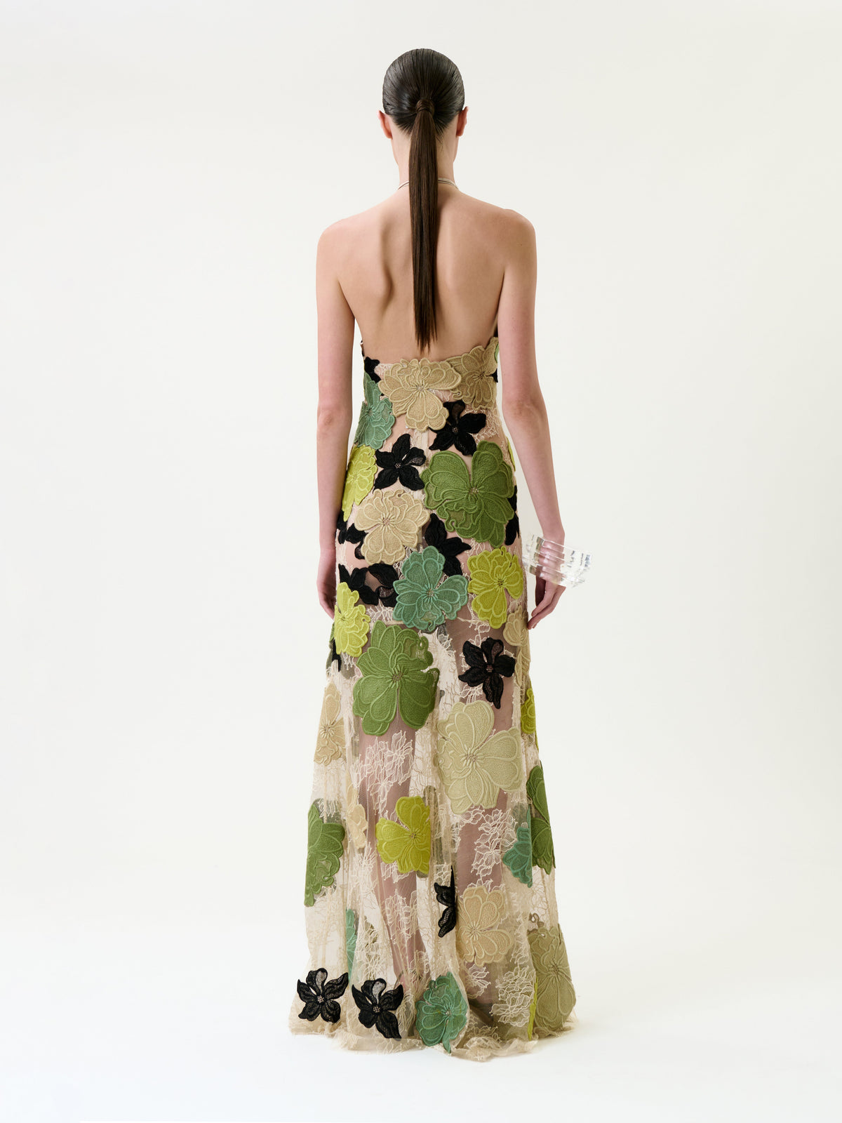 A woman stands facing away in the Xenia Dress, a strapless, floor-length gown featuring bold green, yellow, and black floral patterns. Her straight hair is tied back as she holds a clear clutch in her right hand against a plain white background.