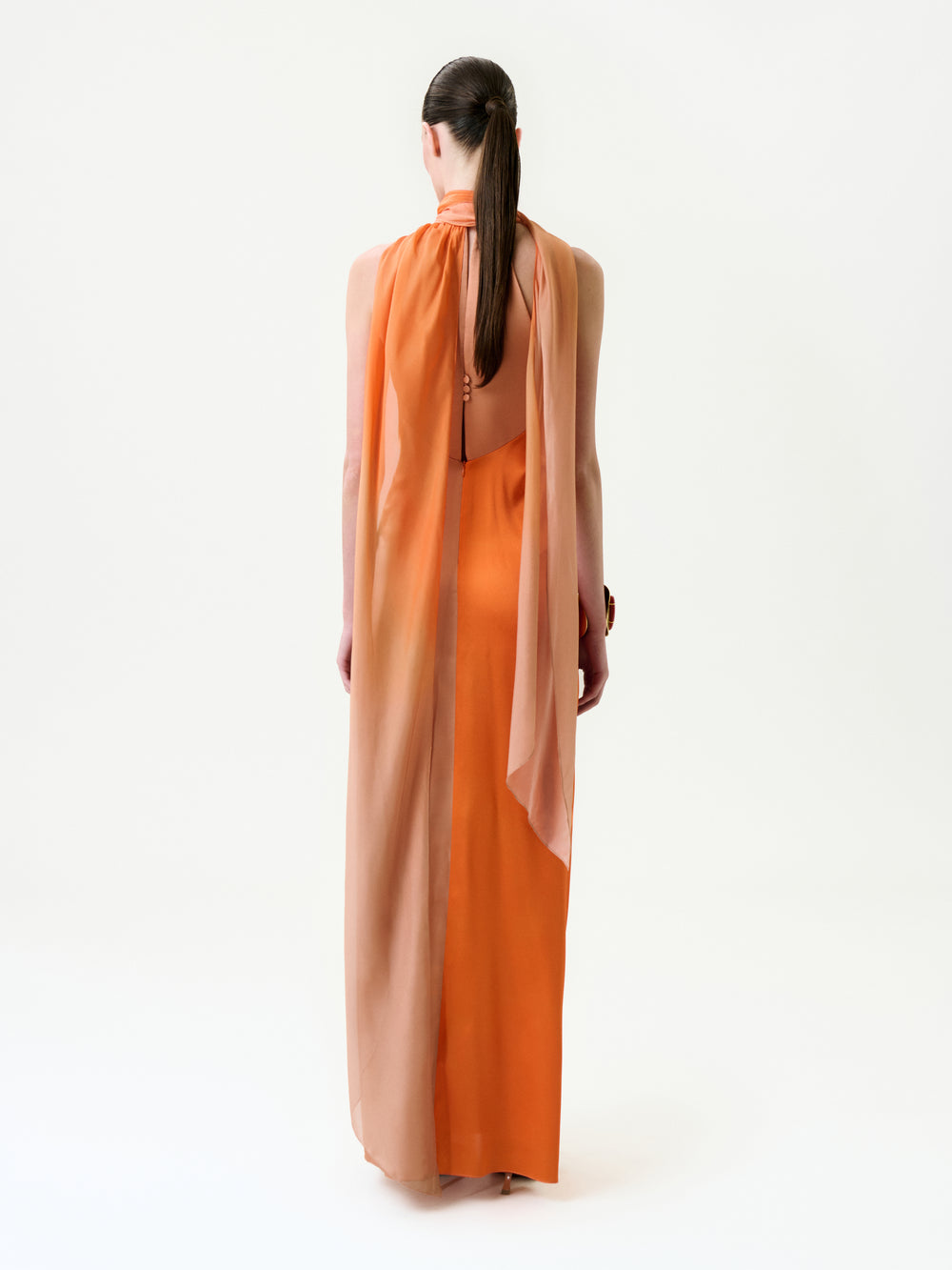 A person with long dark hair tied back is seen from behind, wearing the Margalit Dress—a sleeveless silk maxi with a flowing sheer peach overlay and high neckline—against a plain white background.