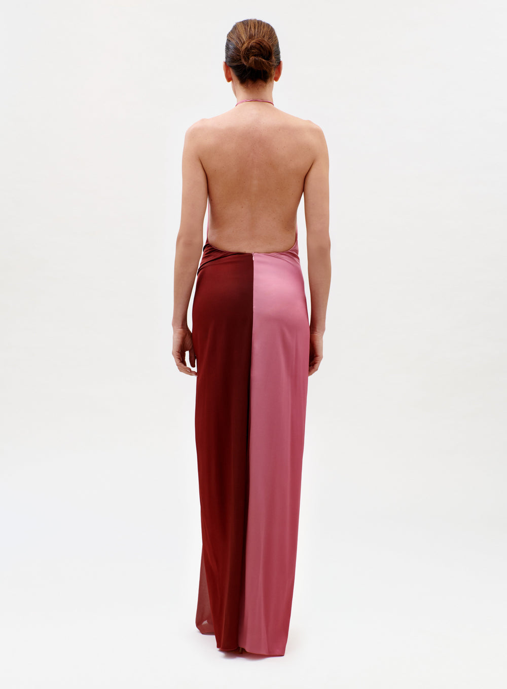 A person with hair in a low bun models the Corsario Dress, a strapless, backless gown featuring vertical burgundy and pink panels, standing against a plain white background.