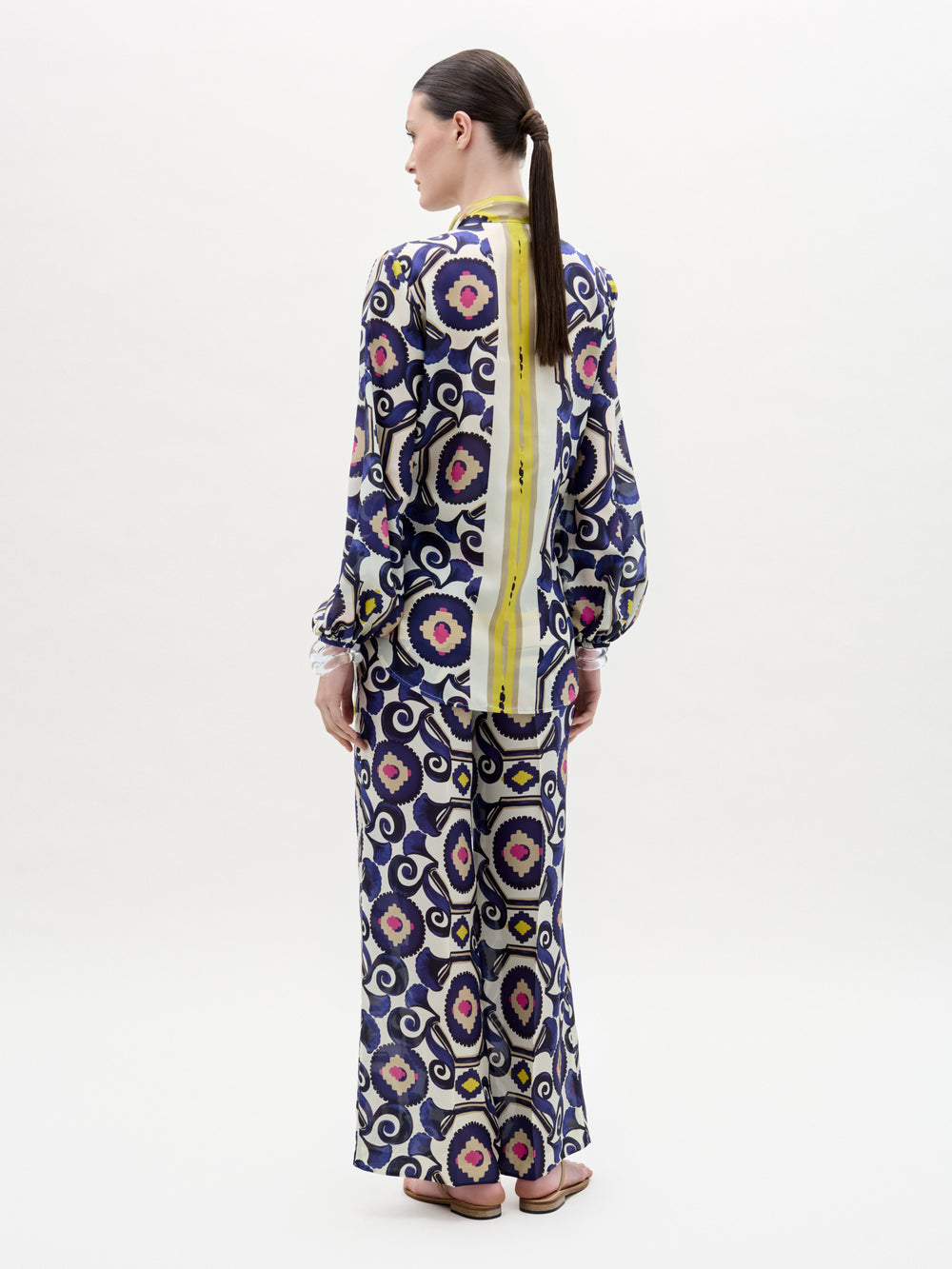 A woman poses against a plain background wearing the Emine Pant—wide-leg silk pants—layered under a long-sleeved, floor-length dress featuring a geometric multicolored pattern in blue, yellow, pink, and white for a chic modern look.