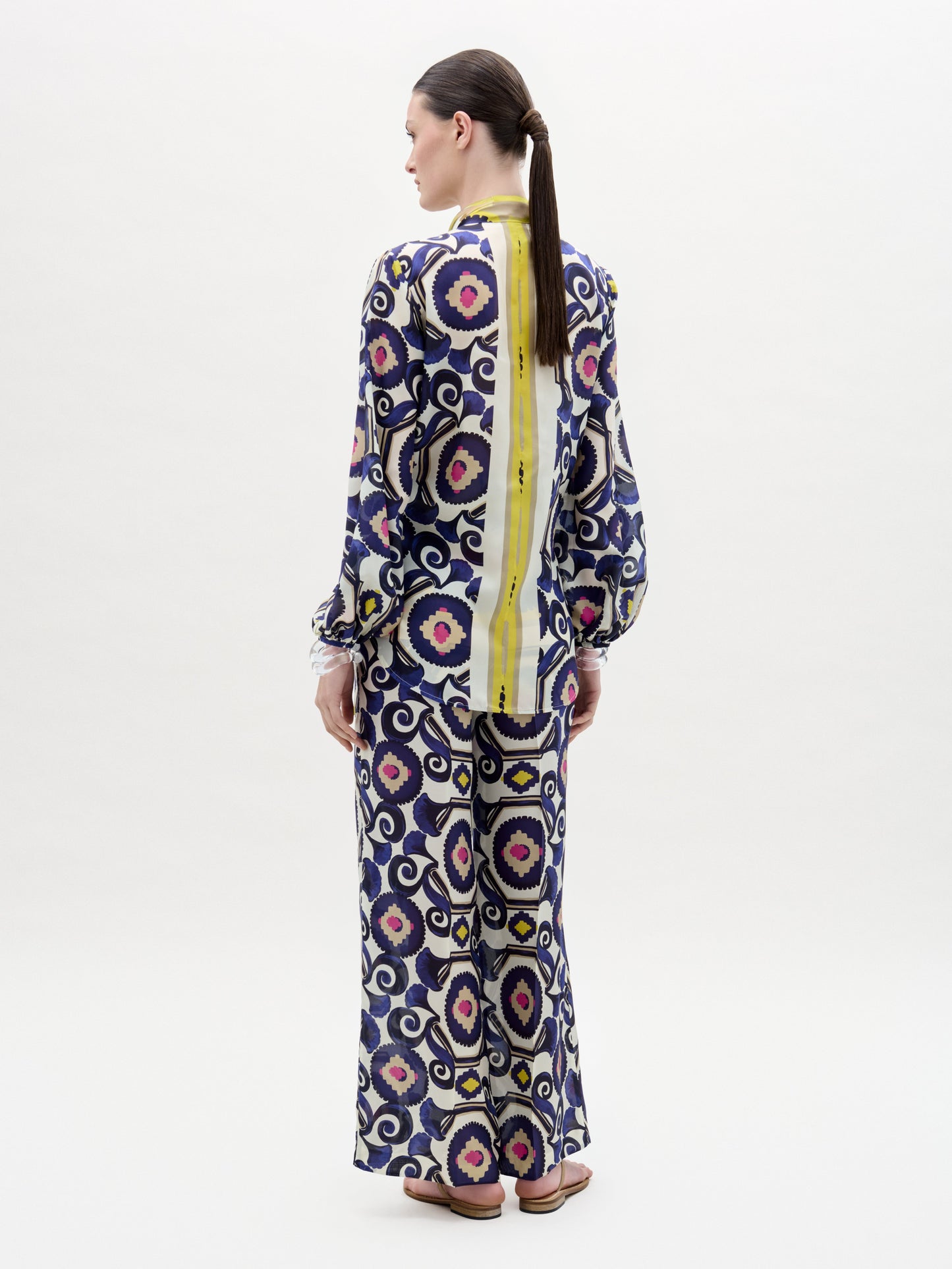 A woman poses against a plain background wearing the Emine Pant—wide-leg silk pants—layered under a long-sleeved, floor-length dress featuring a geometric multicolored pattern in blue, yellow, pink, and white for a chic modern look.