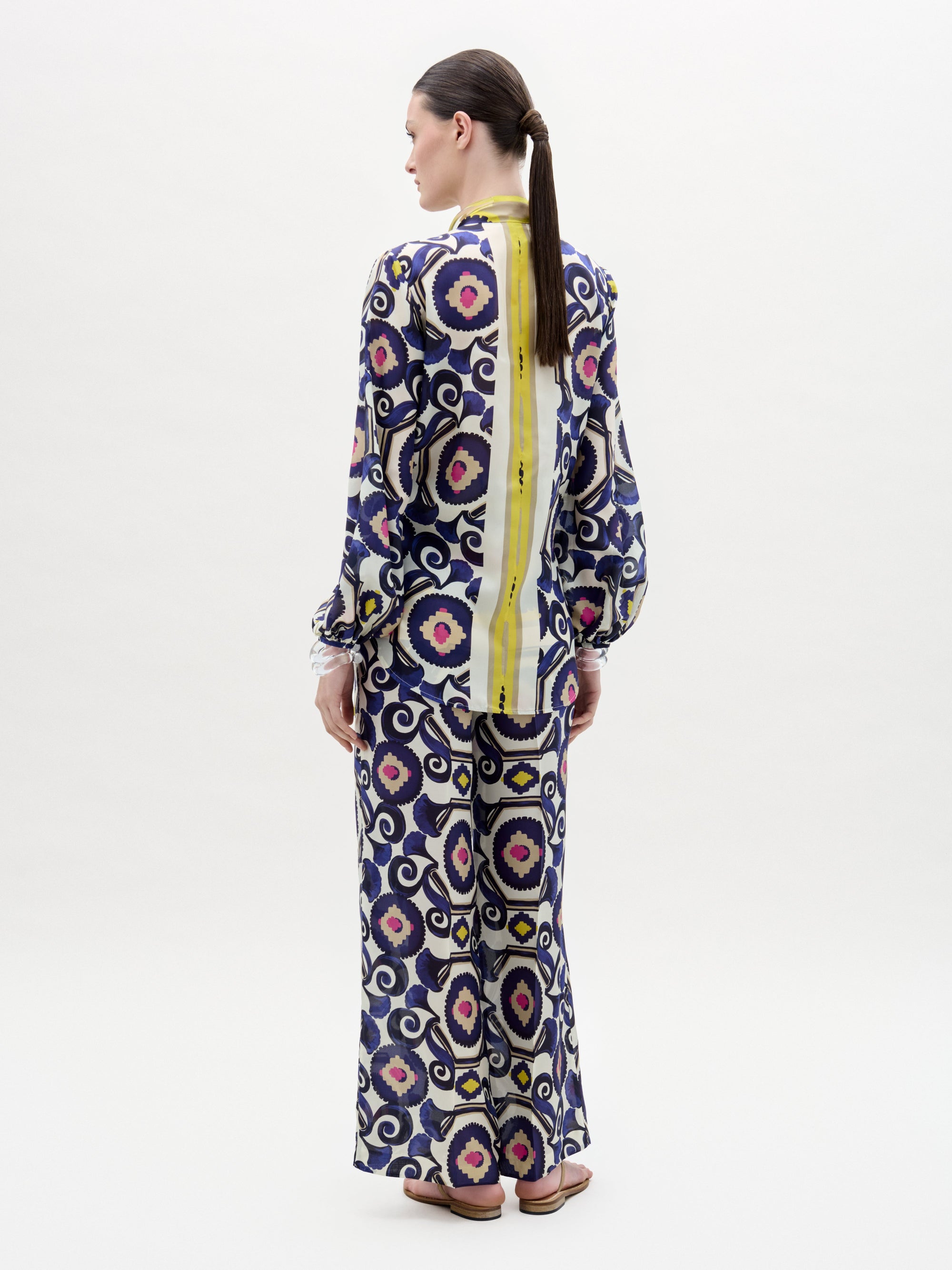 A woman poses against a plain background wearing the Emine Pant—wide-leg silk pants—layered under a long-sleeved, floor-length dress featuring a geometric multicolored pattern in blue, yellow, pink, and white for a chic modern look.