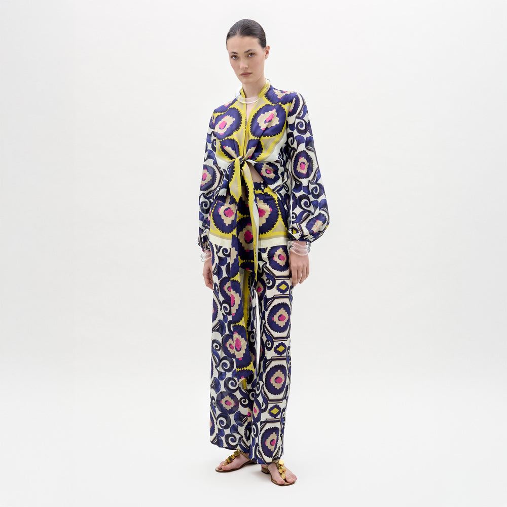 A woman poses against a plain background wearing the Emine Pant—wide-leg silk pants—layered under a long-sleeved, floor-length dress featuring a geometric multicolored pattern in blue, yellow, pink, and white for a chic modern look.
