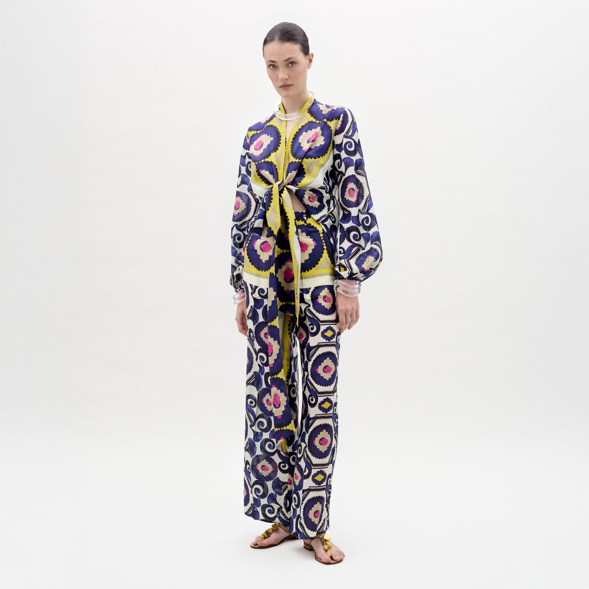 A woman poses against a plain background wearing the Emine Pant—wide-leg silk pants—layered under a long-sleeved, floor-length dress featuring a geometric multicolored pattern in blue, yellow, pink, and white for a chic modern look.