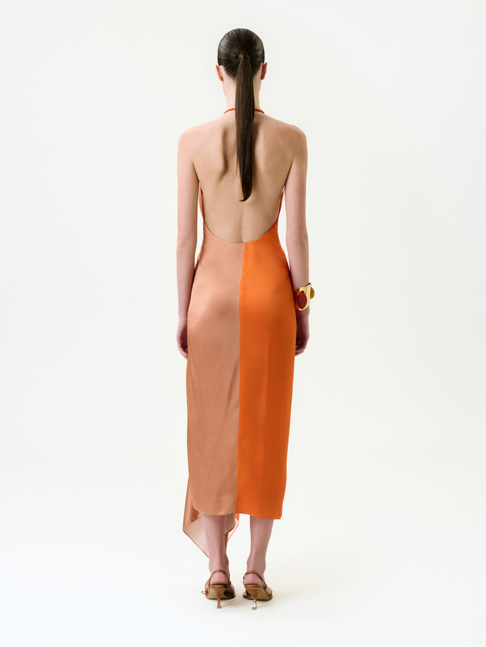 A woman, hair in a low ponytail, models the Syndney Dress—a backless halter-neck maxi in two-tone peach and orange Italian fabric—paired with strappy heels and a chunky bracelet against a plain white background.